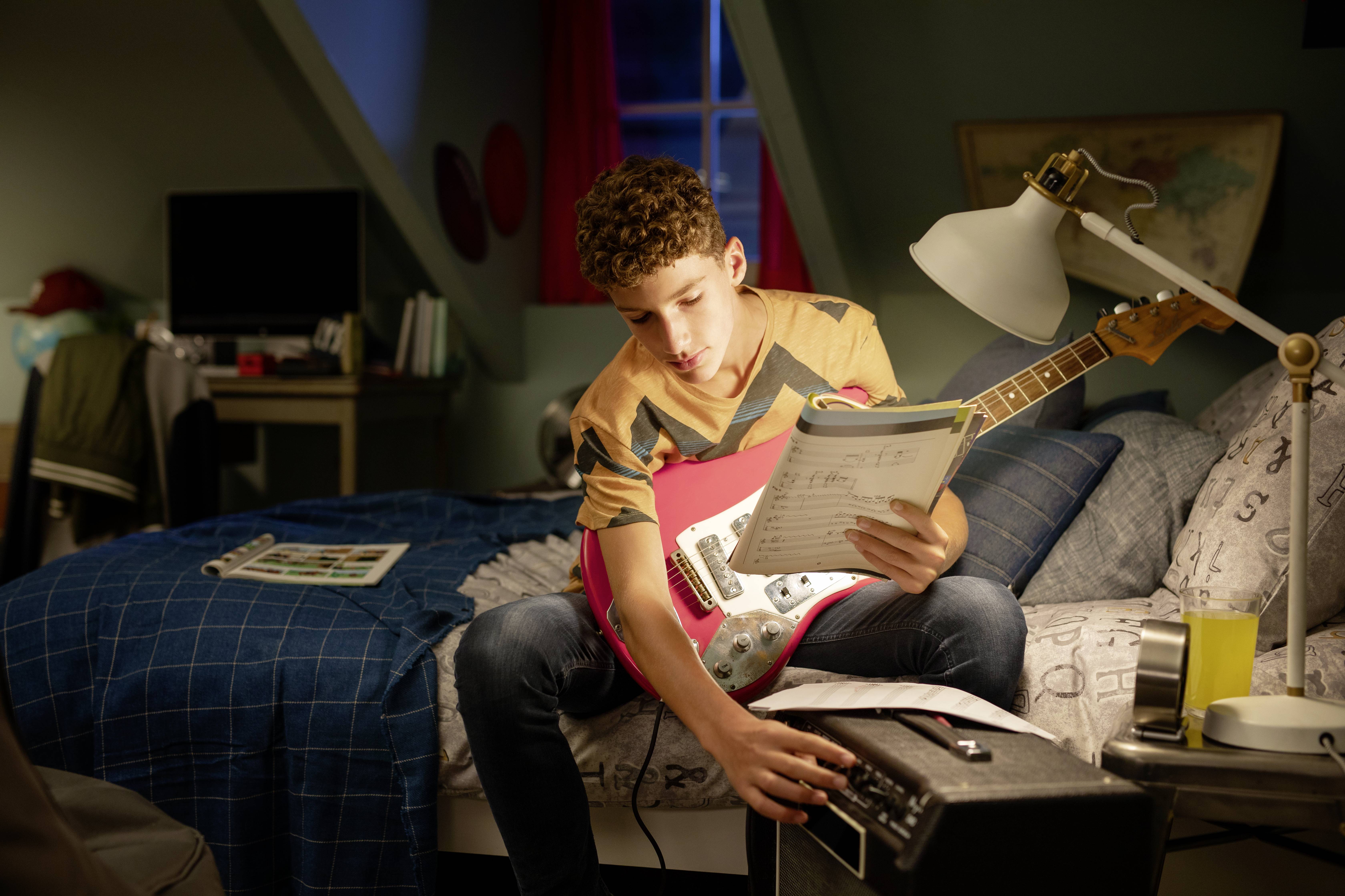 Ein Junge sitzt auf einem Bett, hält eine rote E-Gitarre und studiert Noten, während er einen Verstärker einstellt.