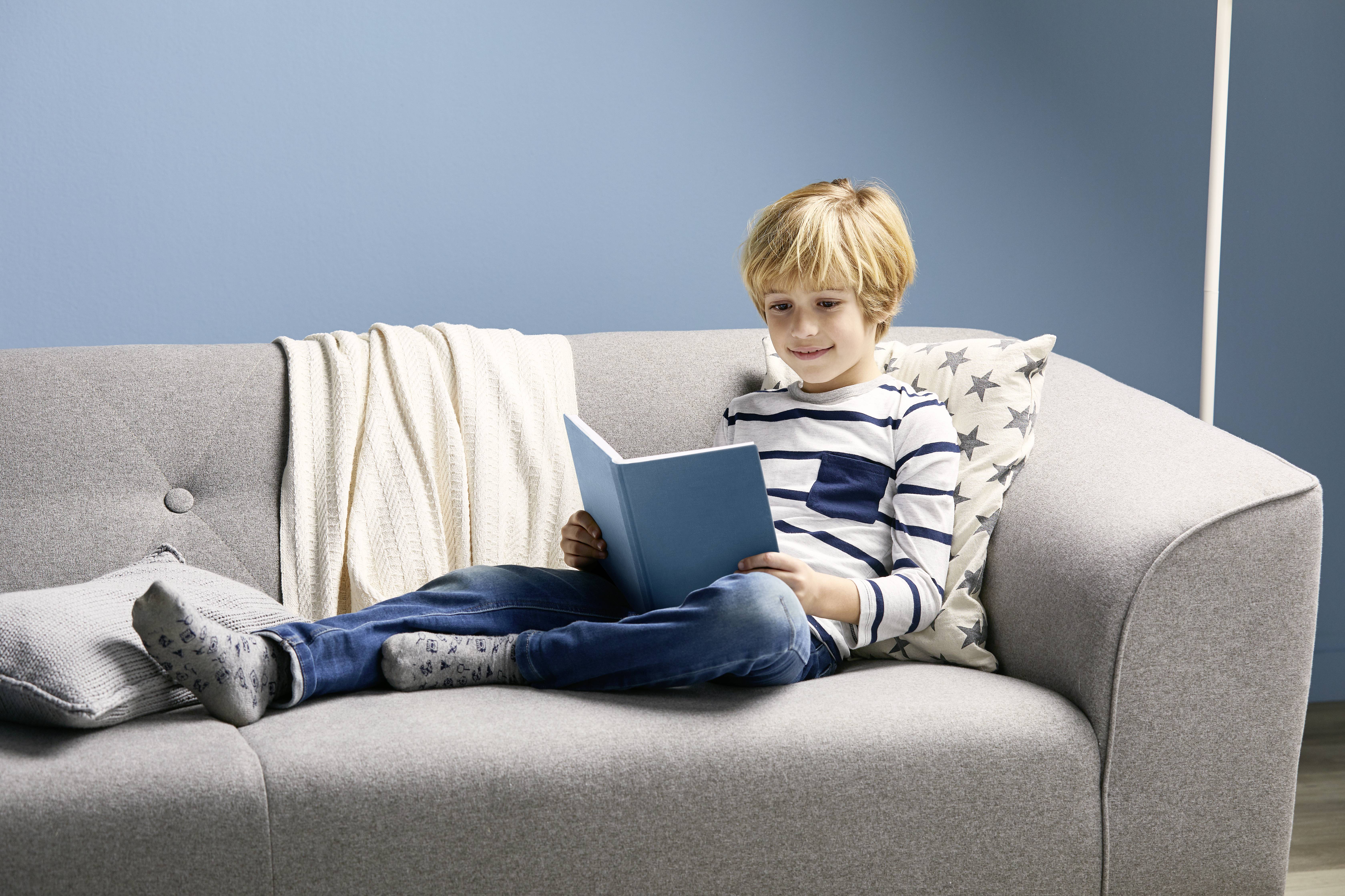 Ein Junge sitzt entspannt auf einem grauen Sofa und liest ein blaues Buch. Neben ihm liegen Kissen und eine Decke.