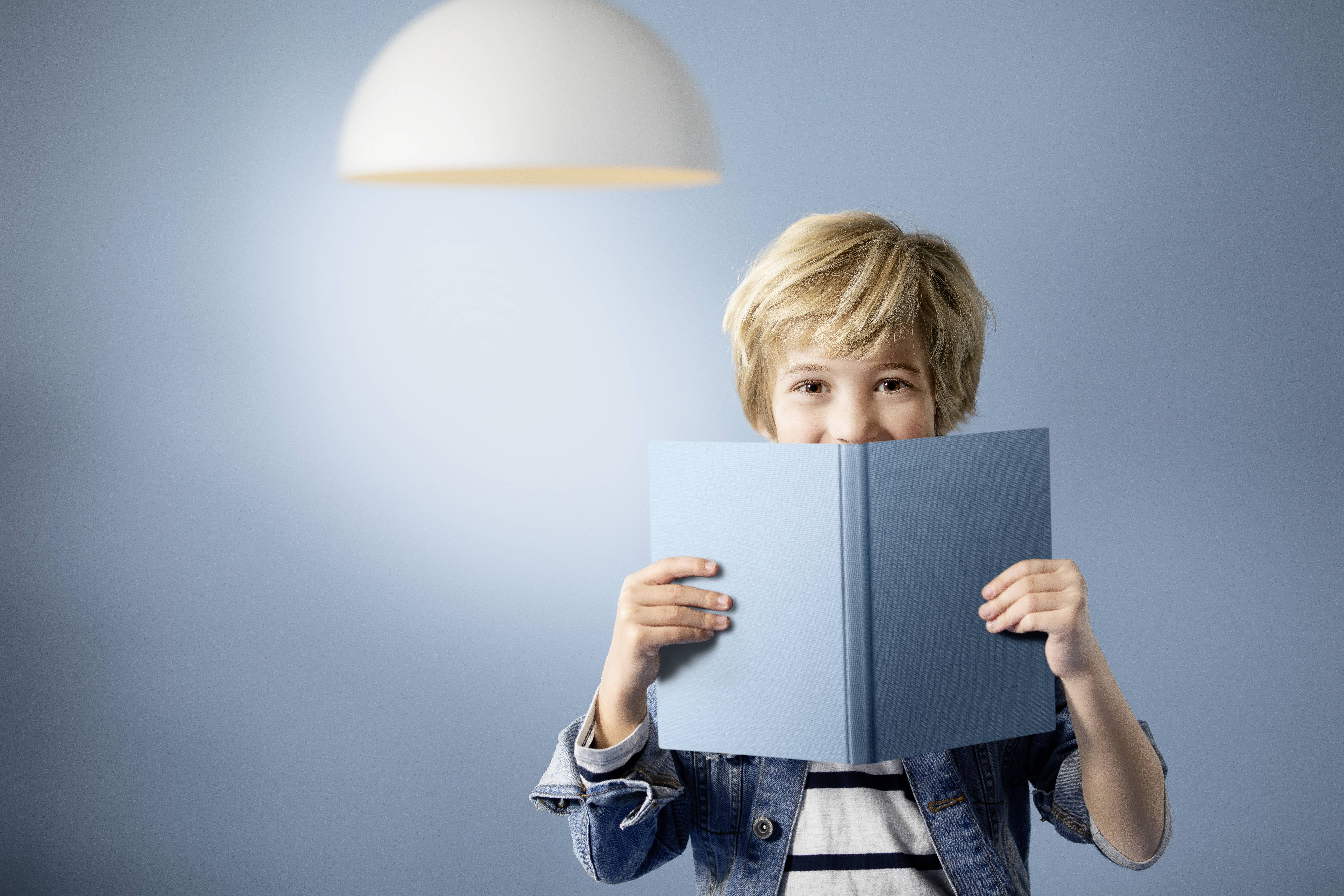 Ein blondes Kind hält ein aufgeschlagenes blaues Buch vor seinem Gesicht, lächelt und steht vor einem hellblauen Hintergrund.