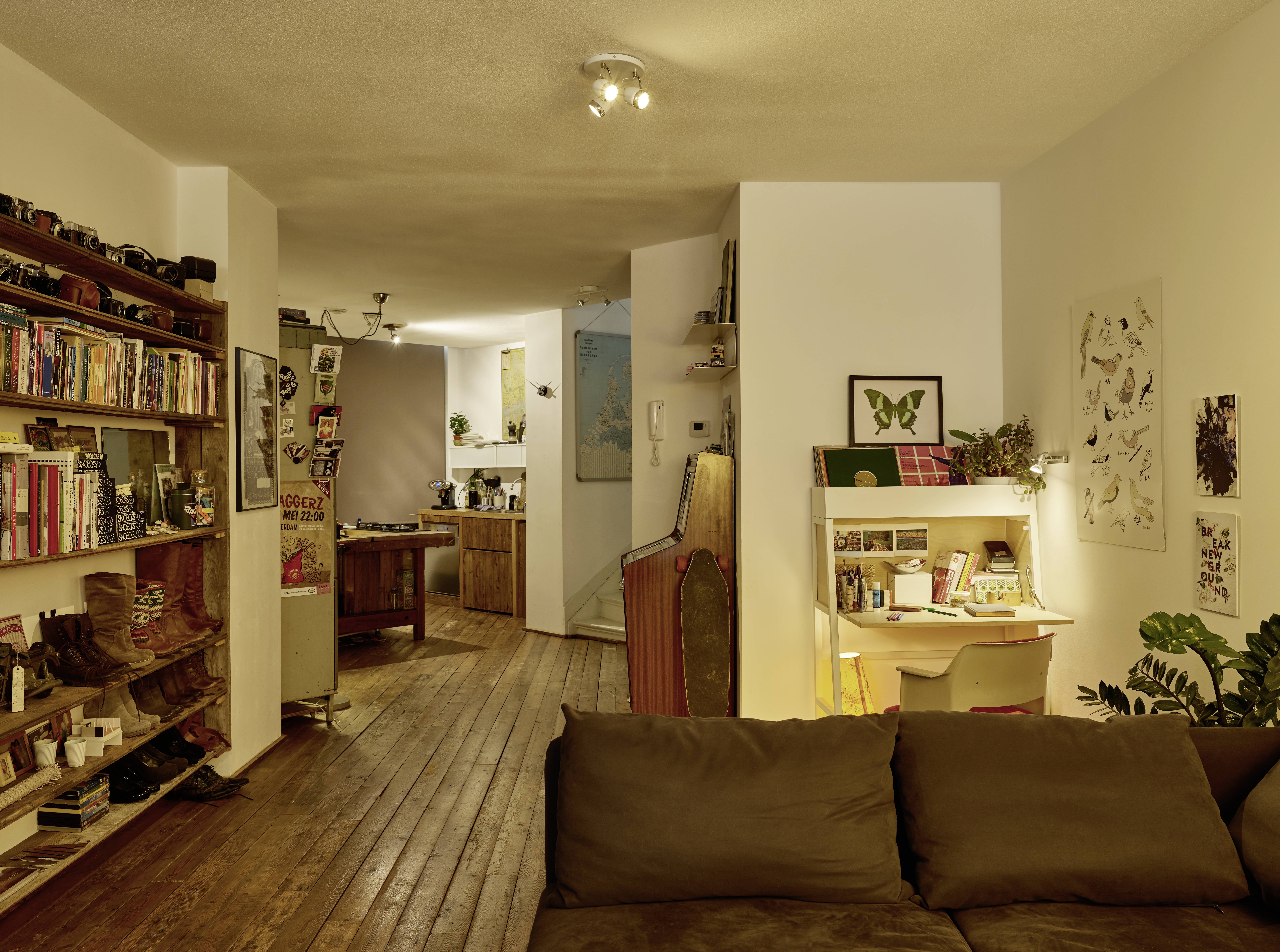 Wohnzimmer mit Holzboden, Bücherregal, Sofa und ein Schreibtisch mit Lampe im Hintergrund. Küche im hinteren Bereich sichtbar.