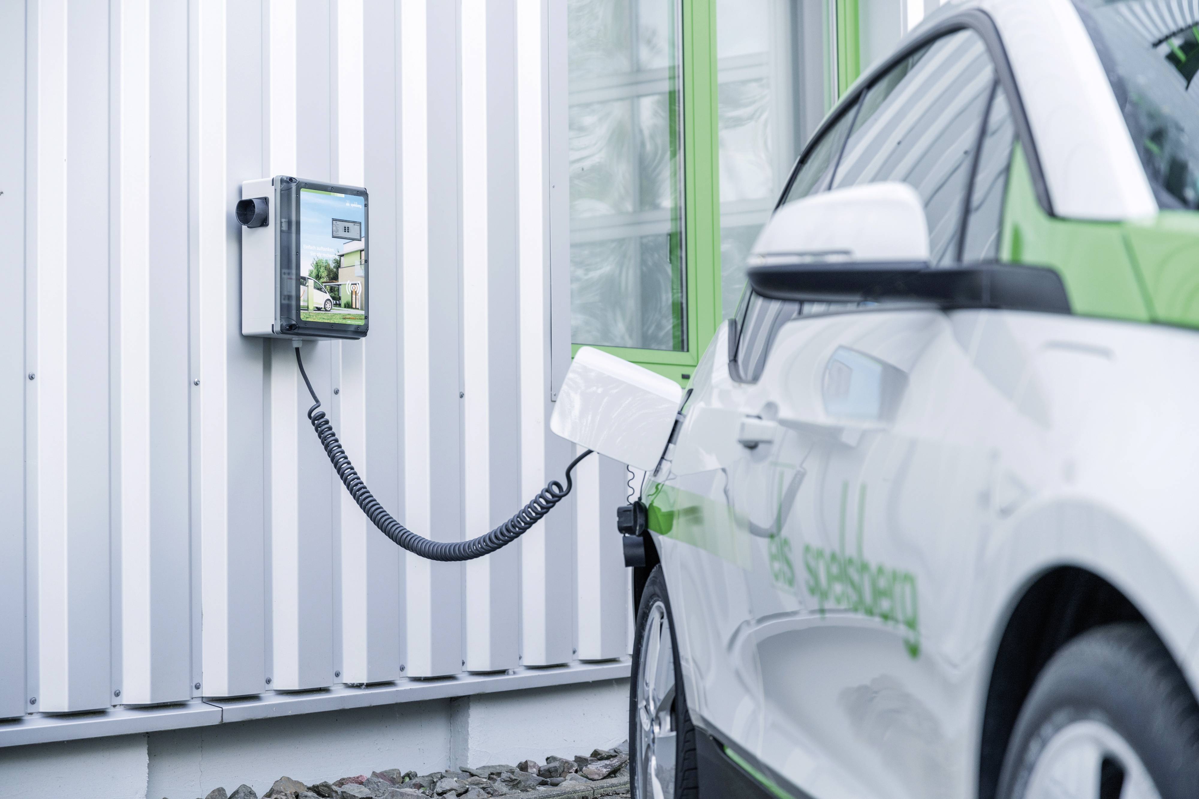 Ein Elektroauto wird an einer Ladestation an der Wand eines Gebäudes aufgeladen. Das Auto ist weiß mit grünen Akzenten.