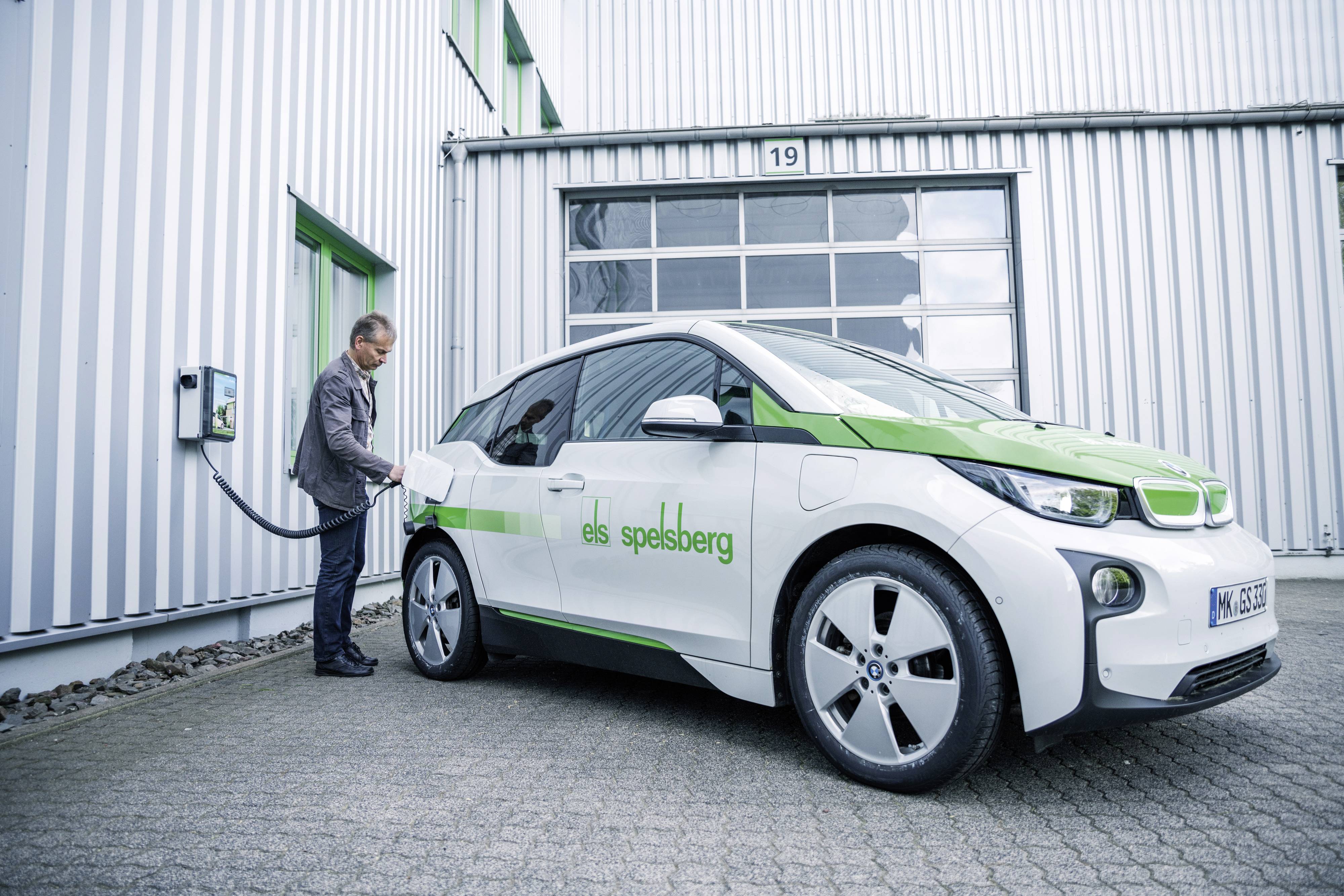 Ein Mann lädt ein weiß-grünes Elektroauto an einer Ladestation vor einem Industriegebäude auf. Das Auto zeigt ein Firmenlogo.
