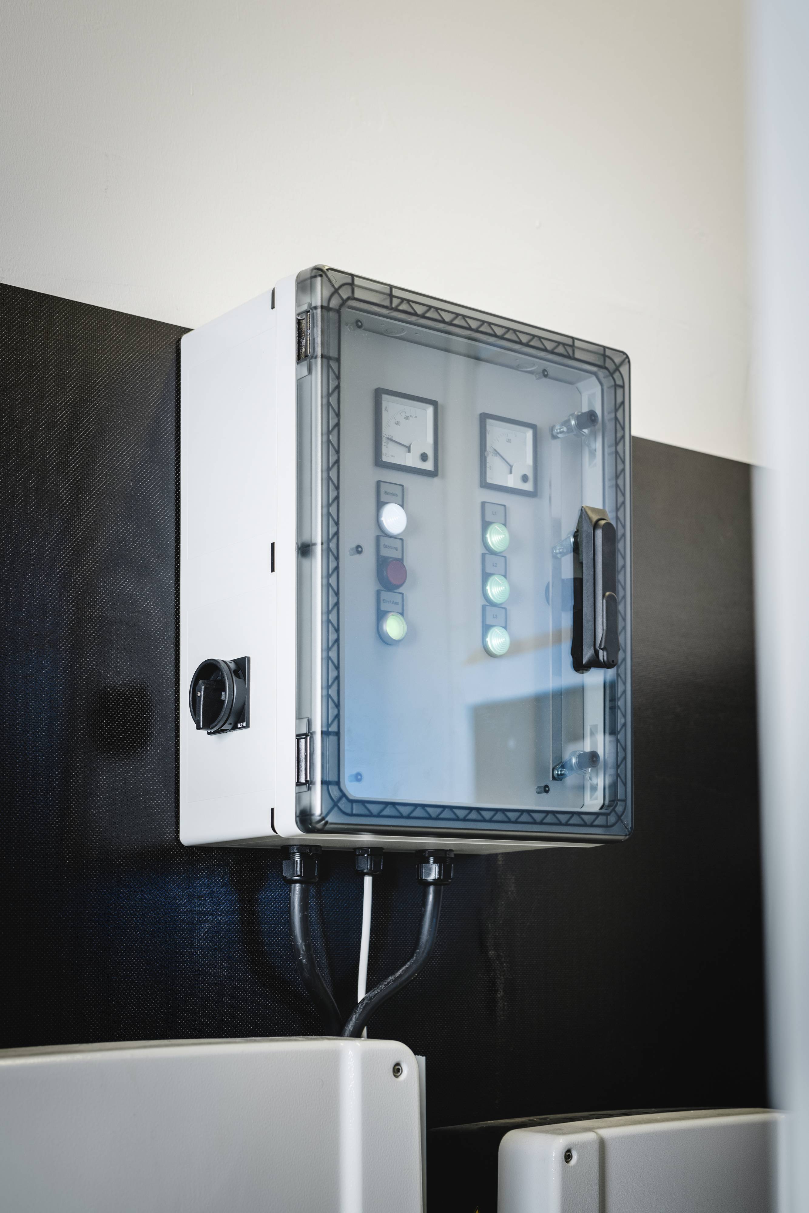 Ein weißer elektrischer Schaltschrank mit transparentem Deckel, mehreren Kontrollleuchten und Schaltern, an einer Wand montiert.