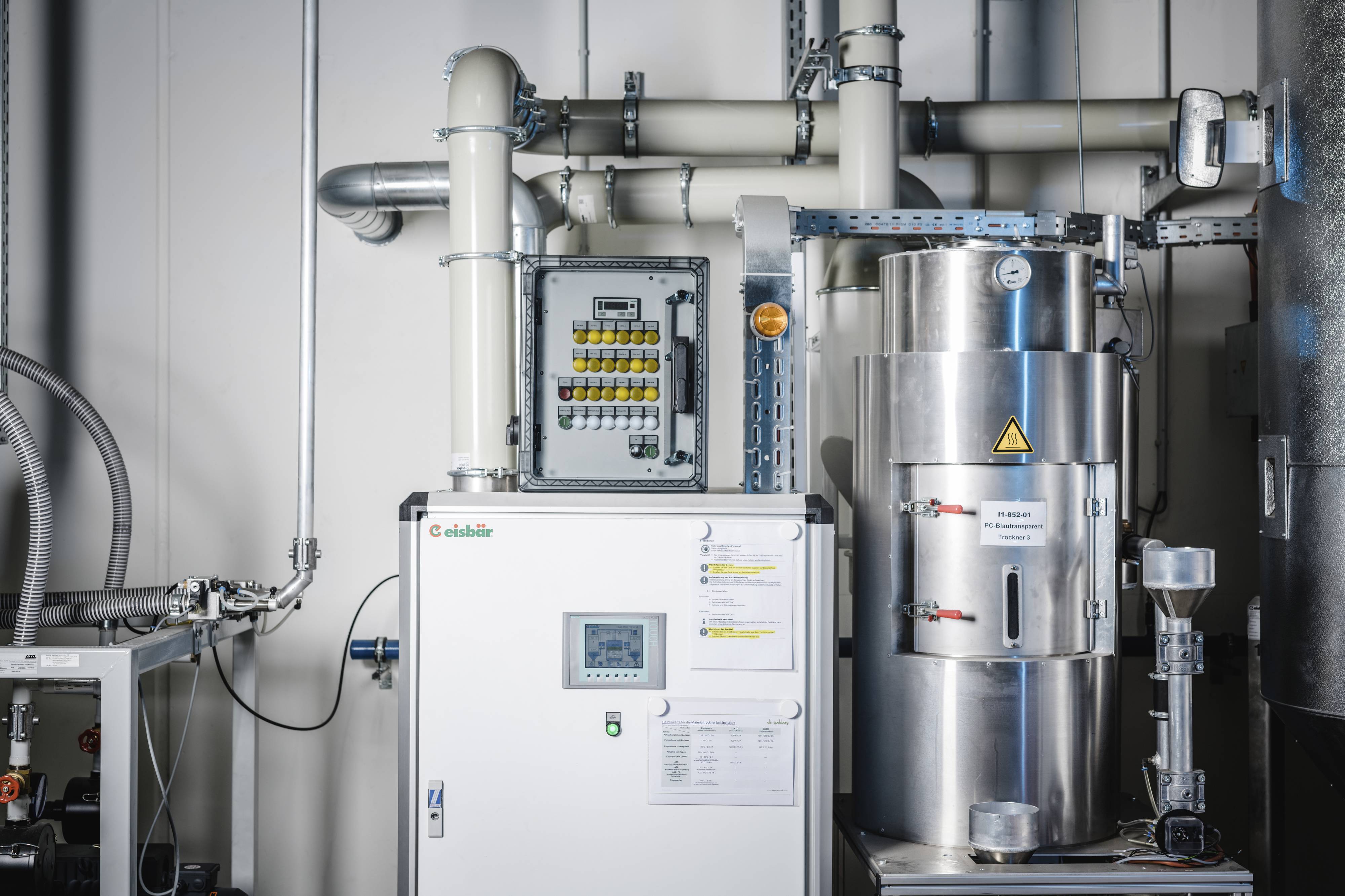 Industrielle Maschine mit Steuerungspanel, Rohren und Metallbehältern in einem technischen Raum. Hauptfunktion: Steuerung und Verarbeitung.