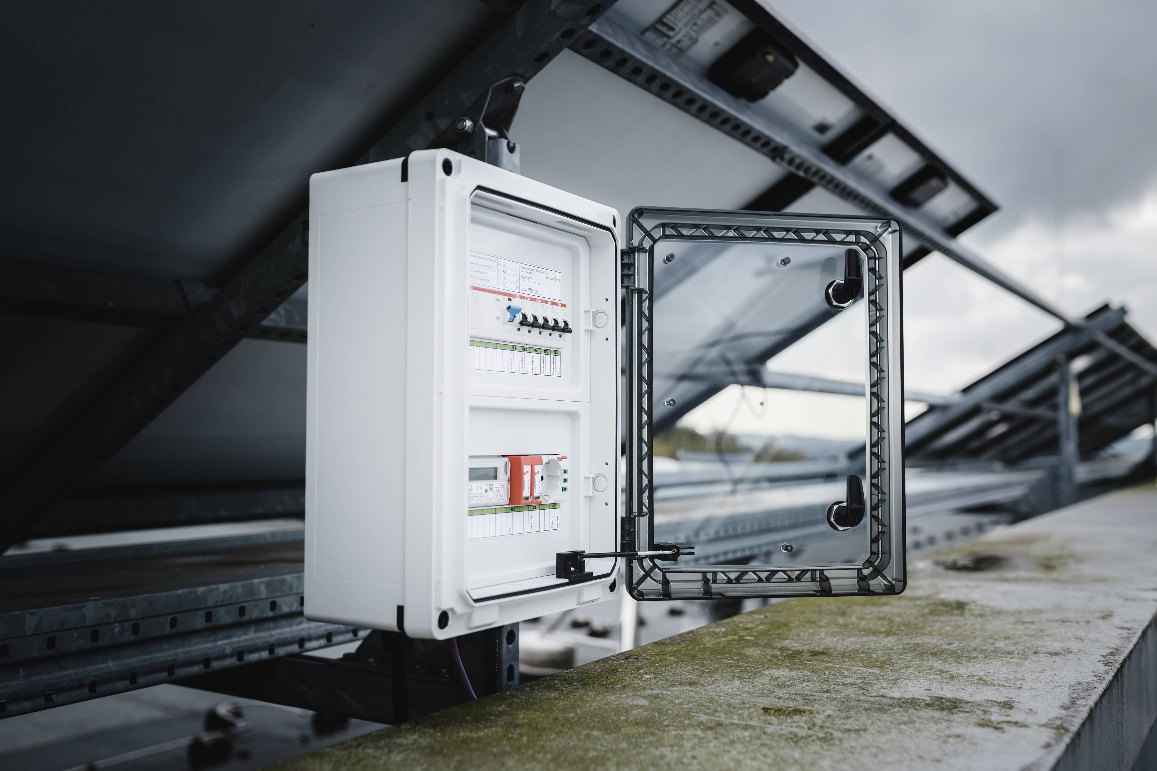 Eine geöffnete elektrische Schalttafel neben Solarmodulen auf einem Dach. Der Himmel ist bewölkt.