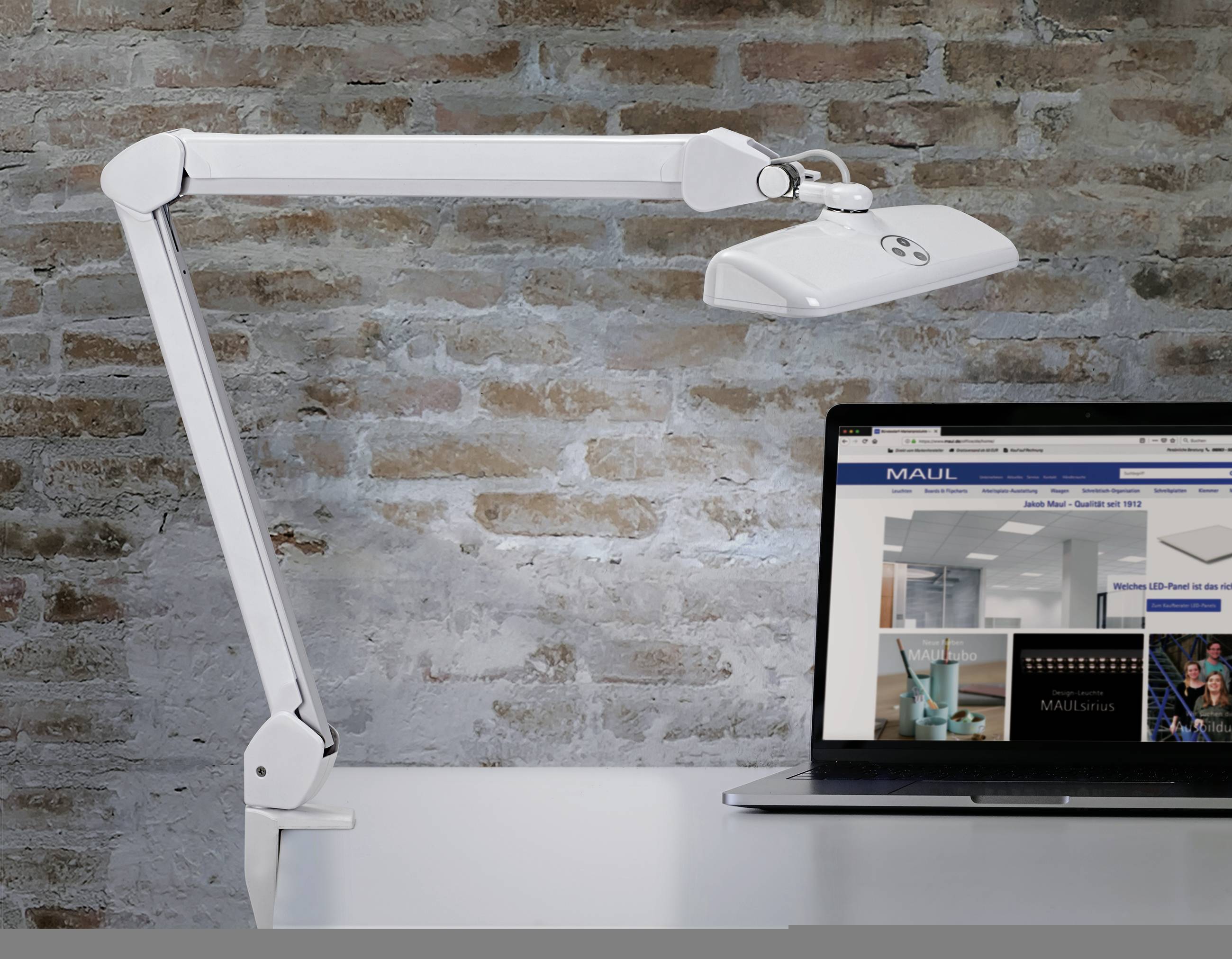 Eine weiße Schreibtischlampe mit Gelenkarm steht auf einem Tisch neben einem Laptop vor einer Backsteinwand. Der Laptop zeigt eine Webseite.