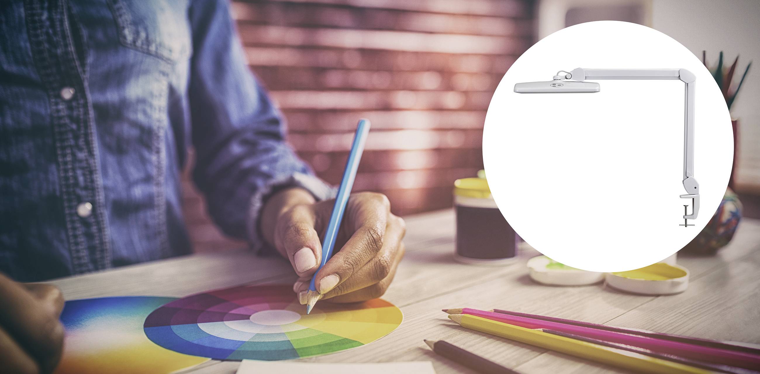 Eine Person malt mit einem Bleistift auf einem Farbkreis. Im Vordergrund ist eine weiße Tischlampe abgebildet.