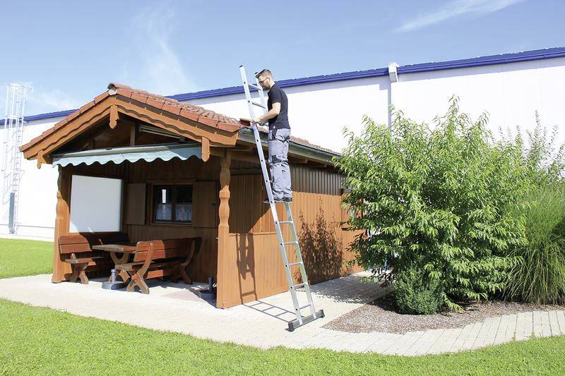 Ein Mann steht auf einer Leiter und repariert das Dach eines kleinen Holzpavillons im Garten. Im Hintergrund ist ein großes Gebäude sichtbar.