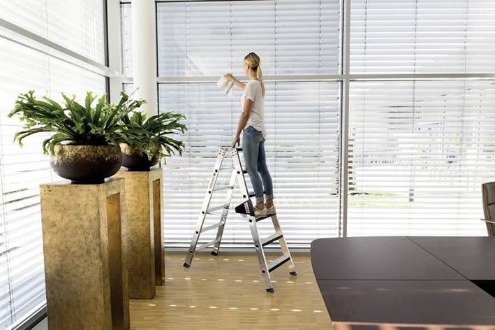 Eine Person steht auf einer Leiter und reinigt große Fensterjalousien. Im Vordergrund befinden sich hohe Pflanzen in Töpfen.