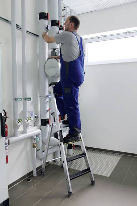 Ein Mann in blauer Arbeitskleidung steht auf einer Leiter und arbeitet an Rohrleitungen in einem technischen Raum mit weißen Wänden.