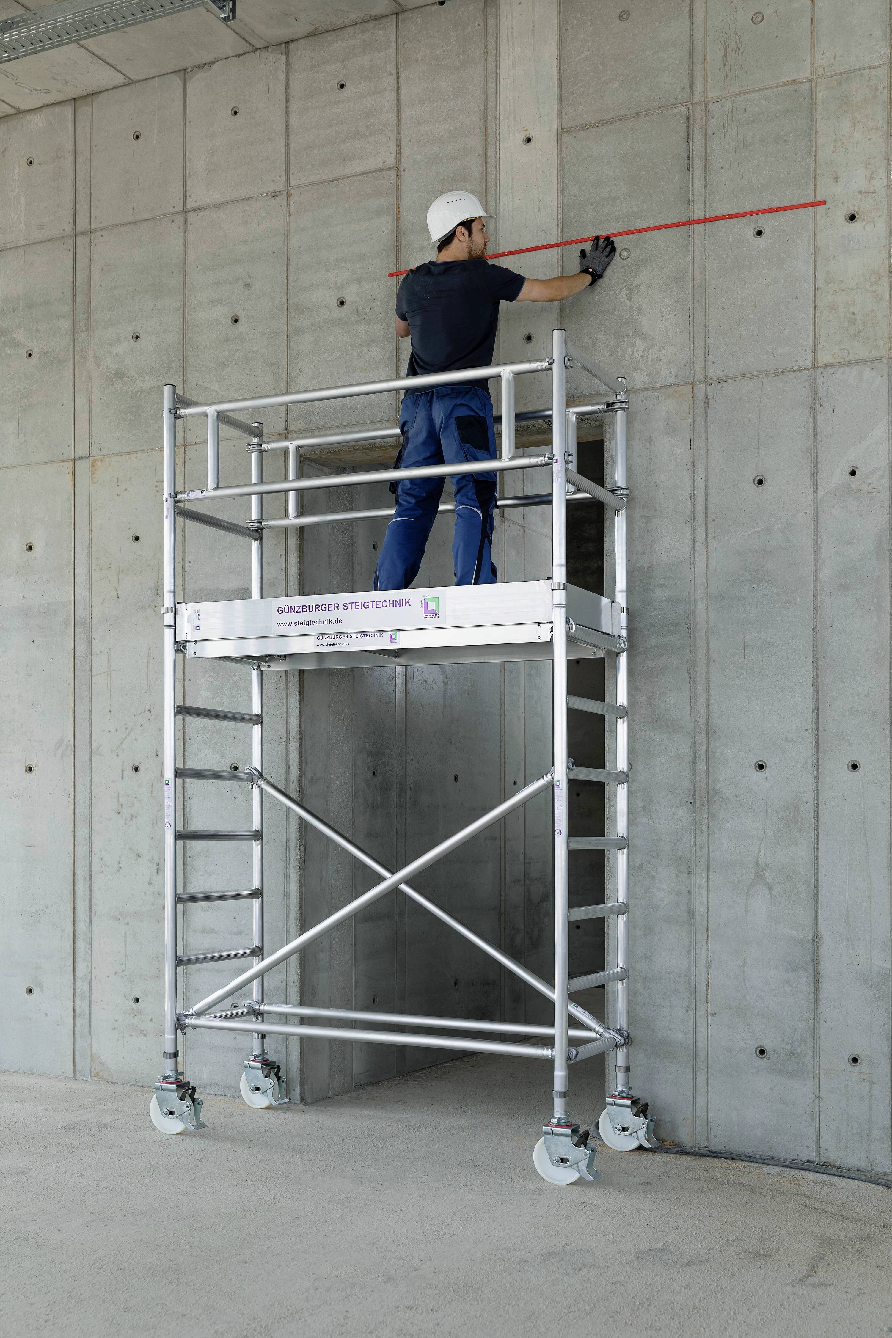Ein Arbeiter auf einem Gerüst misst eine Betonwand mit einem roten Maßband. Er trägt einen Schutzhelm und Arbeitshandschuhe.