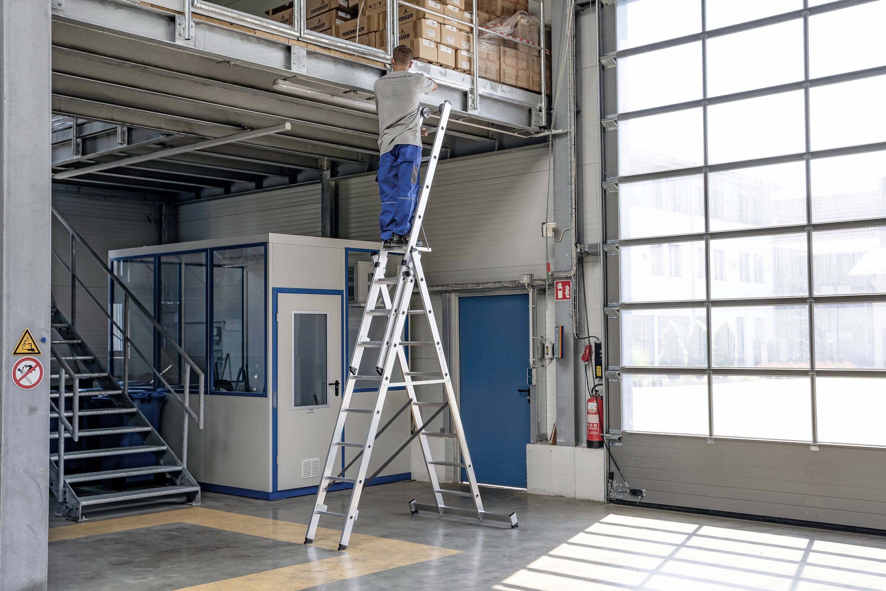 Eine Person steht auf einer hohen Leiter in einem Lagerhaus und greift nach oben zu einem Regal. Es herrscht Tageslicht, rechts sind große Fenster.