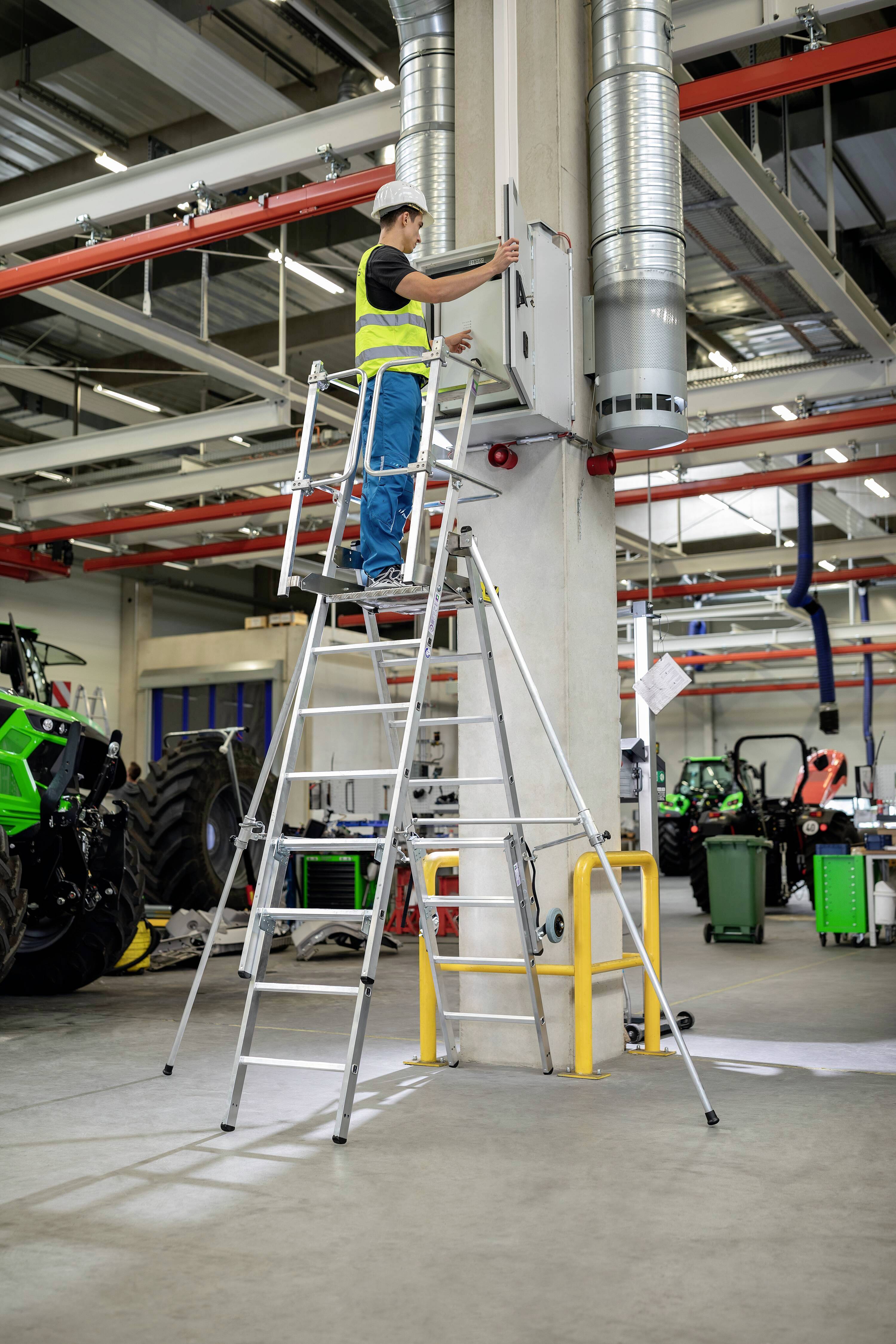 Eine Person steht auf einer Leiter in einer Fabrikhalle und überprüft elektrische Anlagen an der Wand. Im Hintergrund sind Traktoren zu sehen.