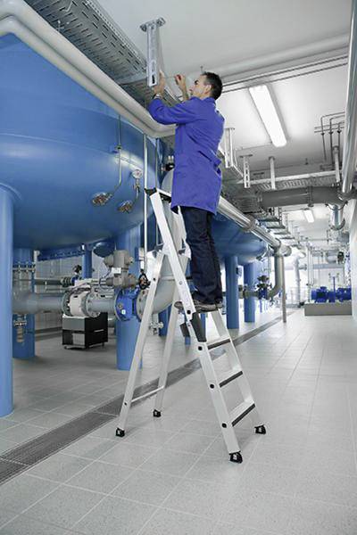 Eine Person in blauer Arbeitskleidung steht auf einer Leiter in einem industriellen Raum und arbeitet an Rohren an der Decke.