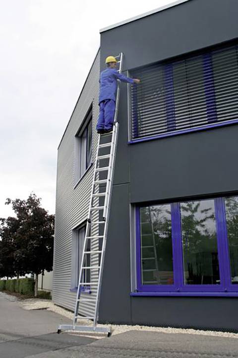 Ein Arbeiter in blauer Schutzkleidung steht auf einer hohen Leiter und reinigt die Fenster eines modernen Gebäude.
