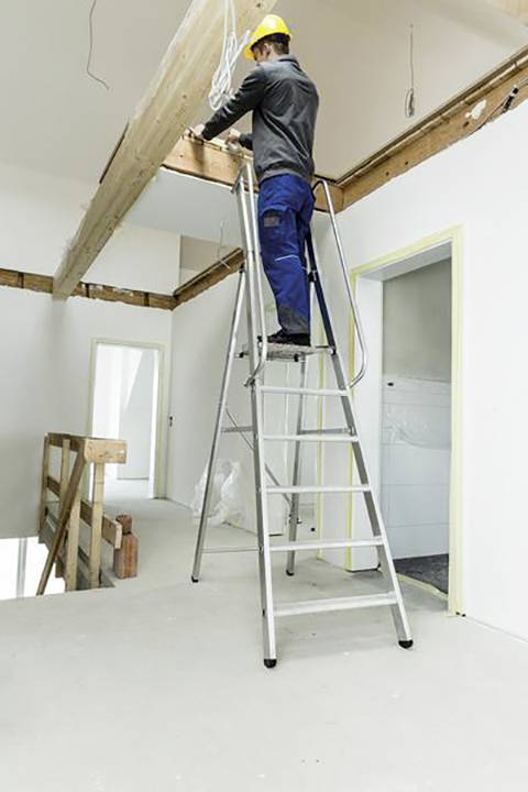 Arbeiter auf Leiter führt elektrische Arbeiten an Holzbalken durch; Baustelle innerhalb eines unvollständigen Gebäudes.