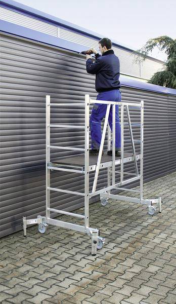 Ein Mann arbeitet mit einem Werkzeug auf einem mobilen Gerüst vor einem geschlossenen Rollladen. Das Gerüst steht auf Pflastersteinen.