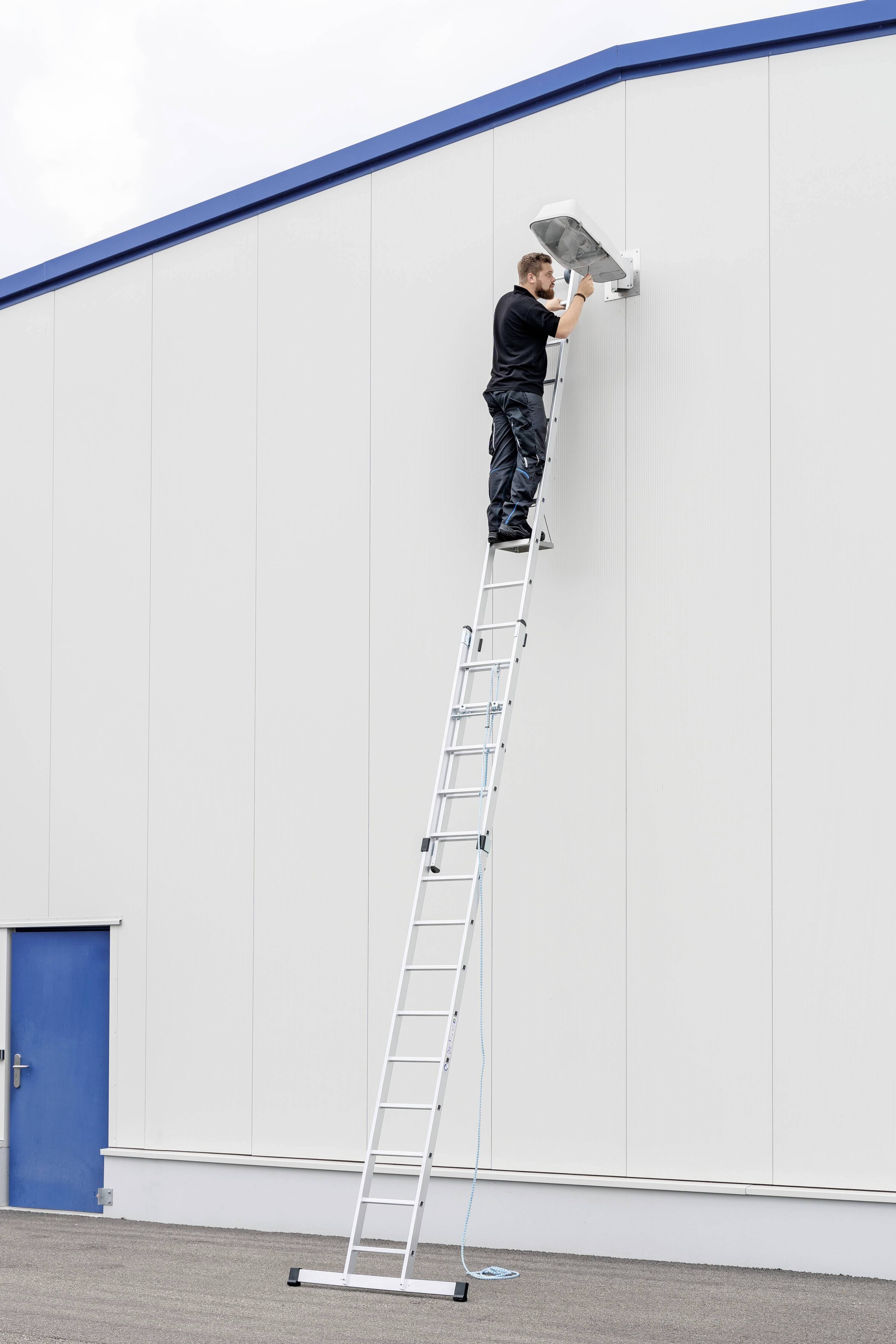 Eine Person steht auf einer hohen Leiter, um an einer Überwachungskamera an der Wand eines großen Gebäudes zu arbeiten.