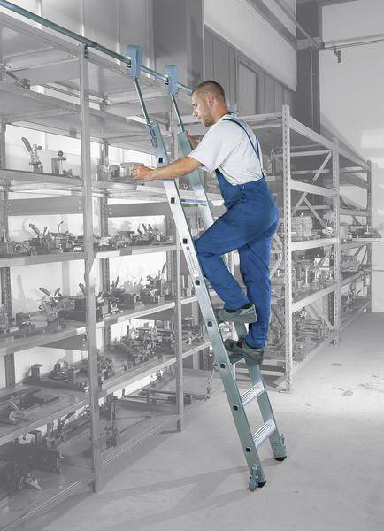 Ein Mann in blauer Arbeitskleidung steht auf einer Leiter und erreicht ein Regal in einem Lagerraum.