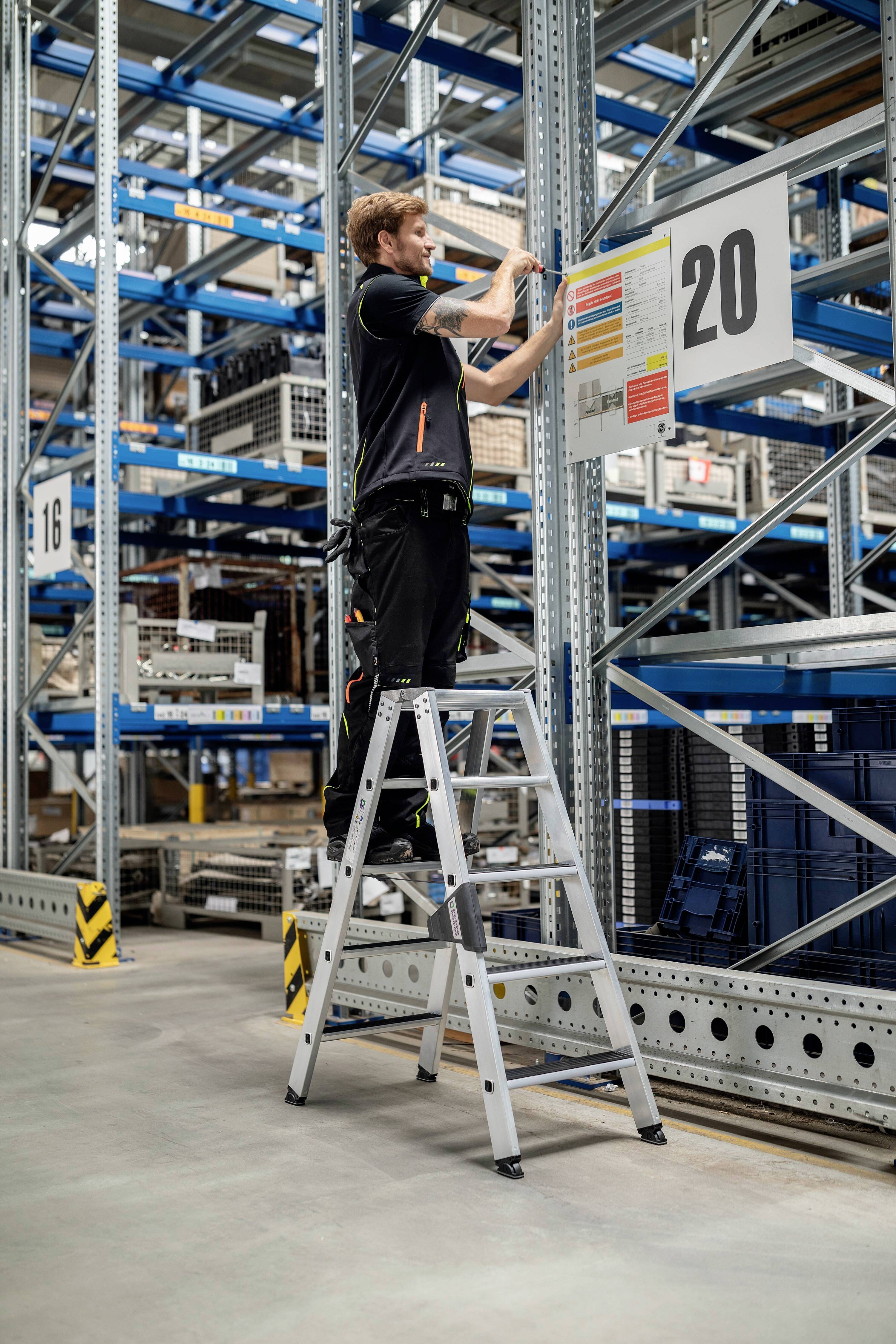 Ein Arbeiter steht auf einer Leiter in einem Lager und befestigt ein Schild mit der Nummer '20' an einem Metallregal.