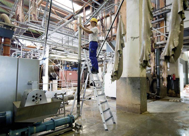 Ein Arbeiter steht auf einer Aluminiumleiter in einer Industriehalle, umgeben von Maschinen und Rohren. Er trägt einen Schutzhelm.