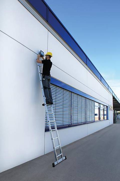 Eine Person mit Schutzhelm arbeitet auf einer Leiter an der Außenwand eines modernen Gebäudes unter blauem Himmel.