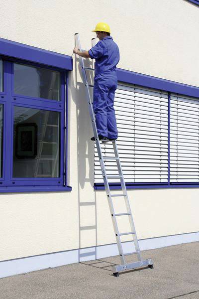 Ein Arbeiter auf einer Leiter, trägt einen gelben Schutzhelm und blaue Arbeitskleidung, überprüft oder repariert etwas an einem Fenster.