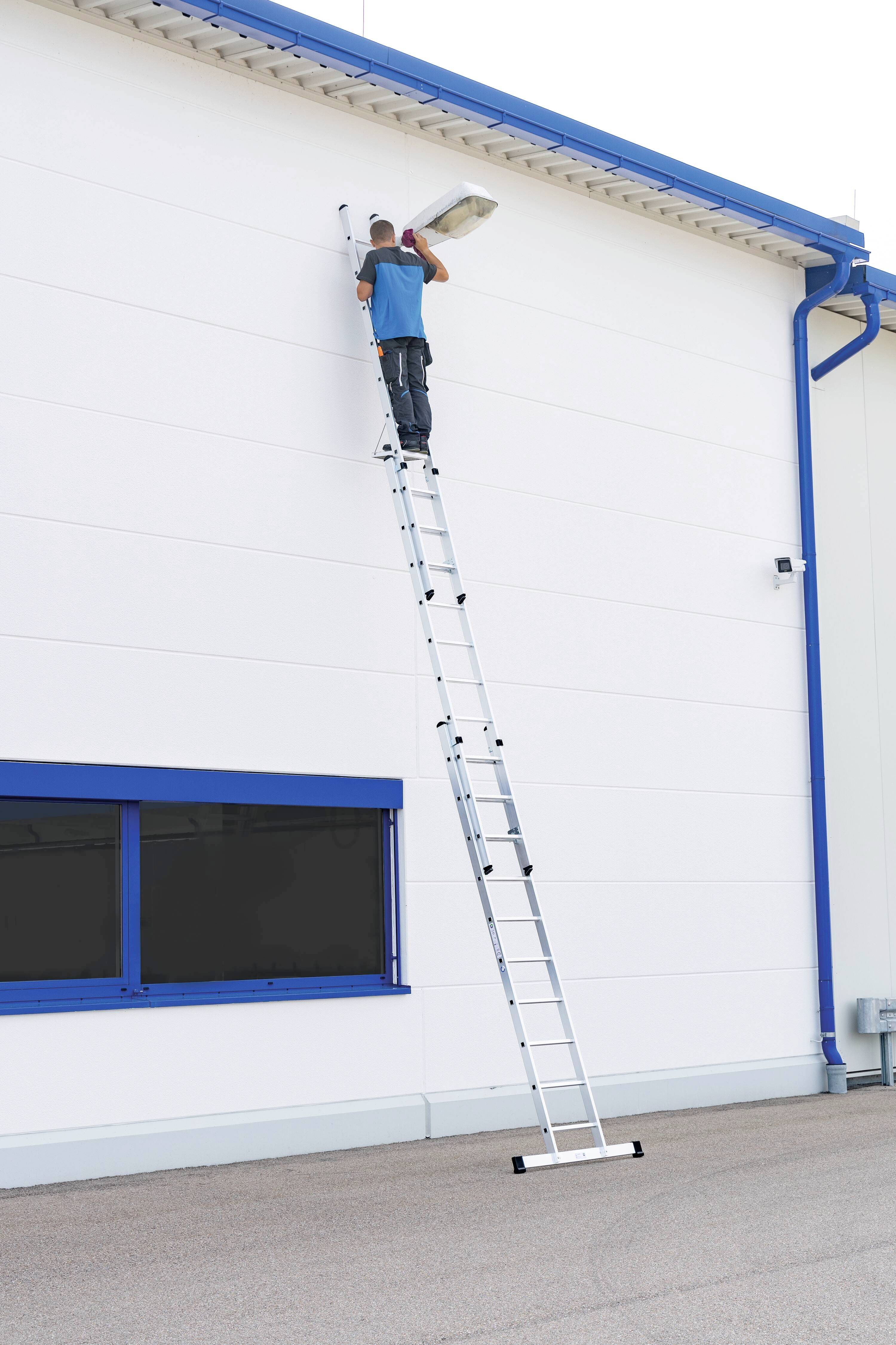 Ein Mann steht oben auf einer langen Leiter und montiert ein Licht an der Wand eines Gebäudes. Die Fenster haben blaue Rahmen.