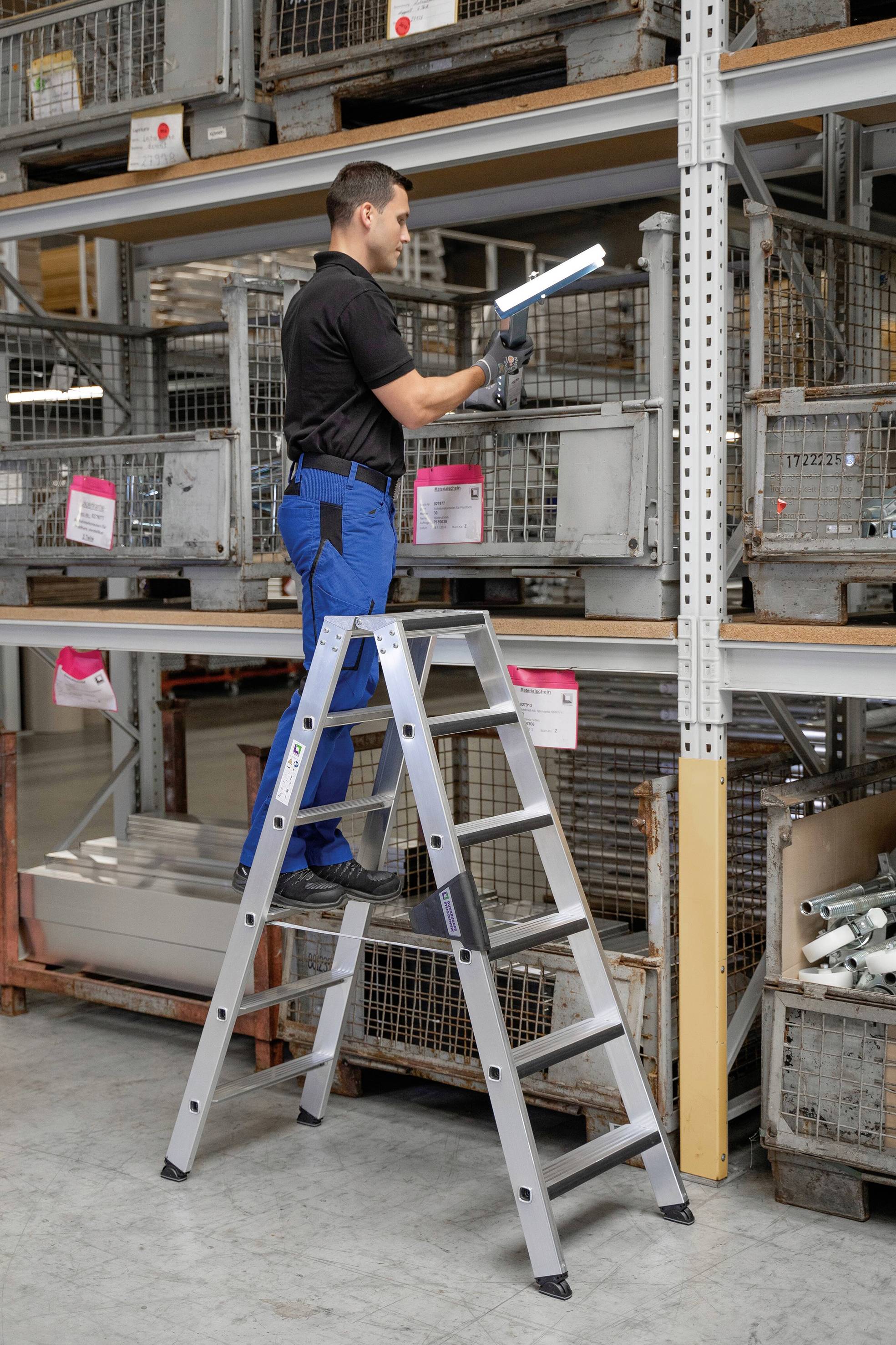 Ein Mann steht auf einer Leiter in einem Lager und überprüft Waren in Regalen. Er trägt Sicherheitskleidung und nutzt ein Tablet.
