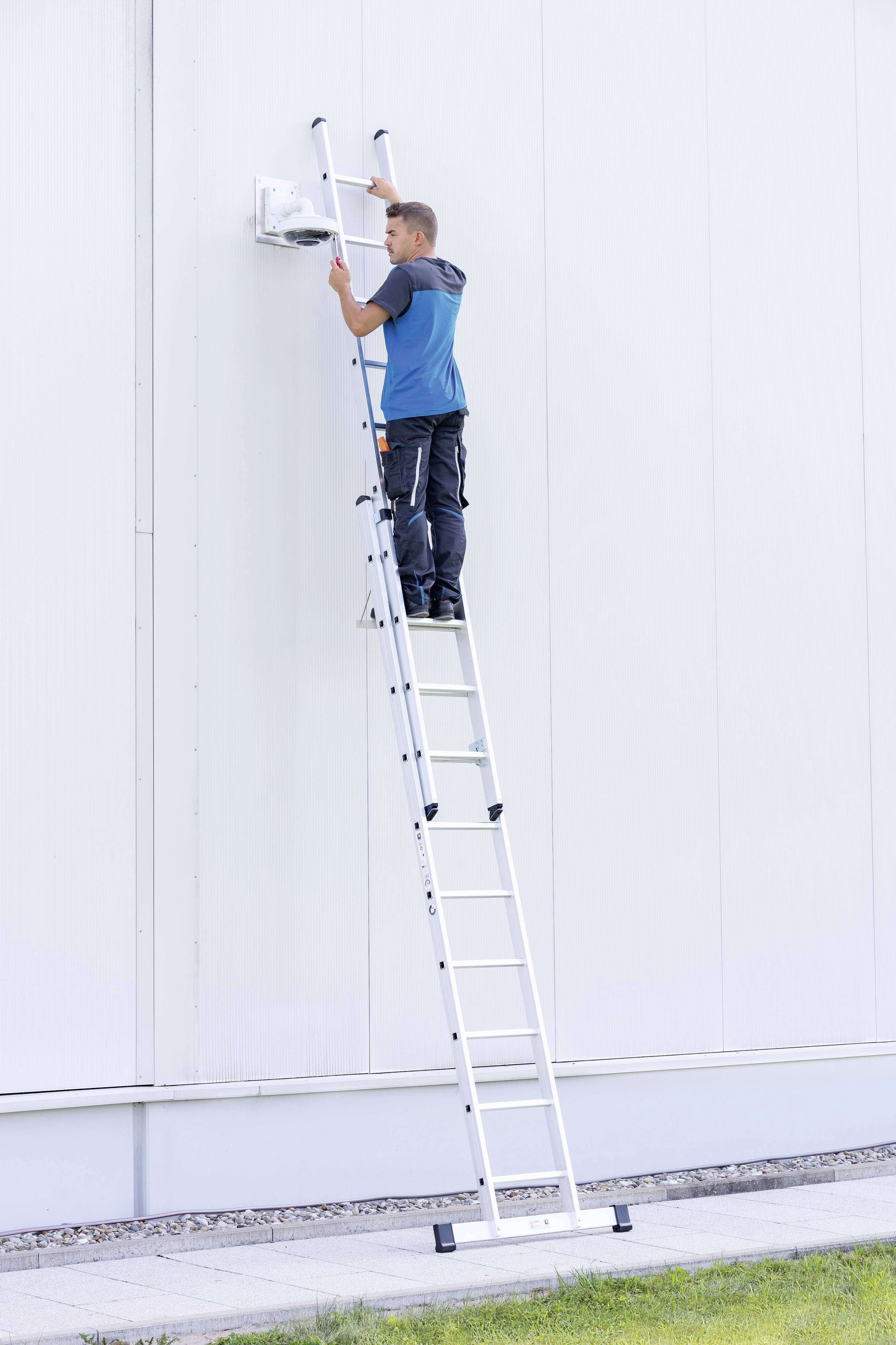 Ein Mann steht auf einer Leiter und arbeitet an einer Wandlampe an einer großen weißen Wand, vermutlich für Wartungsarbeiten.