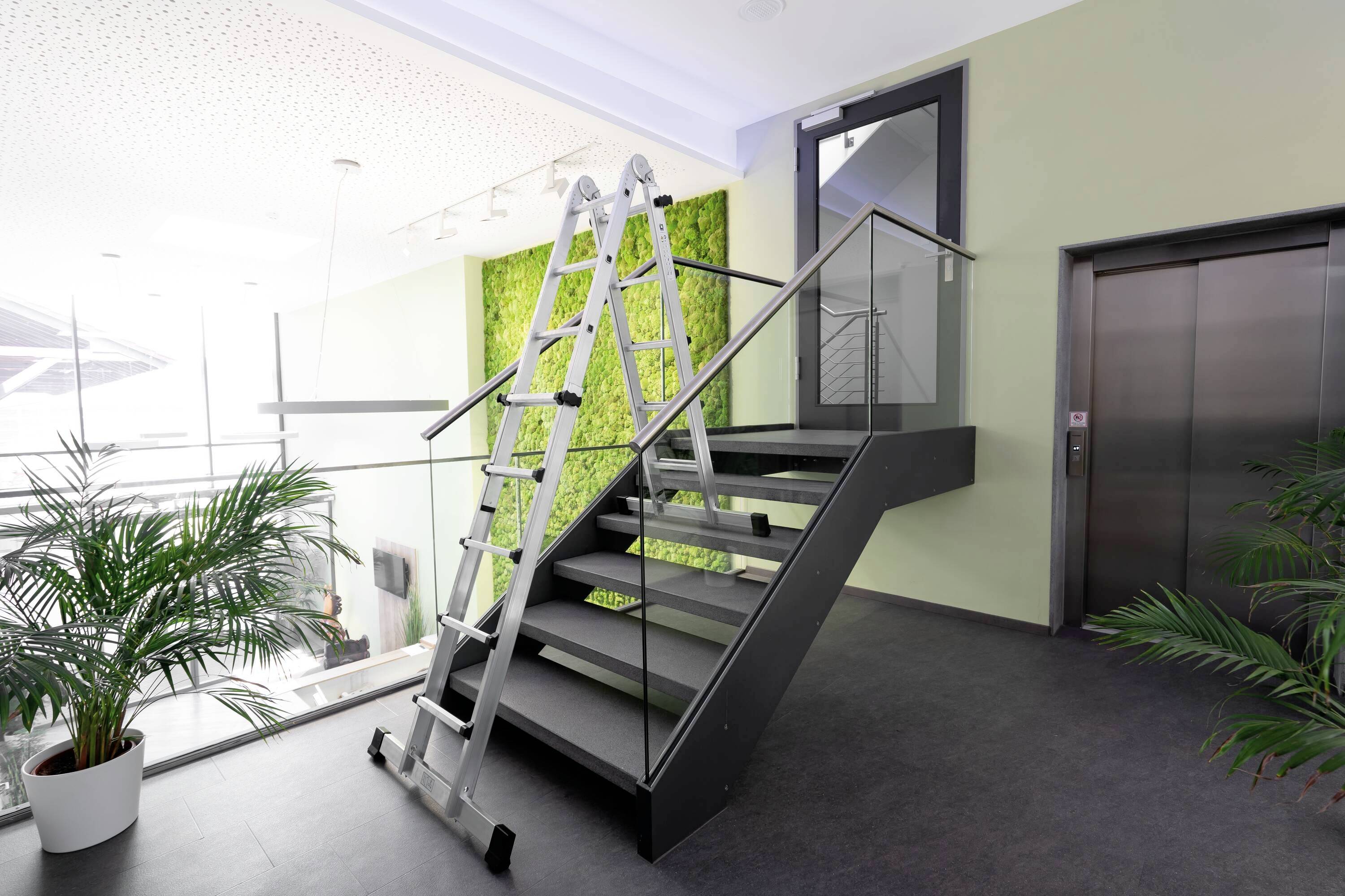 Eine Leiter steht auf einer Treppe in einem modernen Gebäude mit Glasgeländer, Pflanzen und einem Aufzug im Hintergrund.