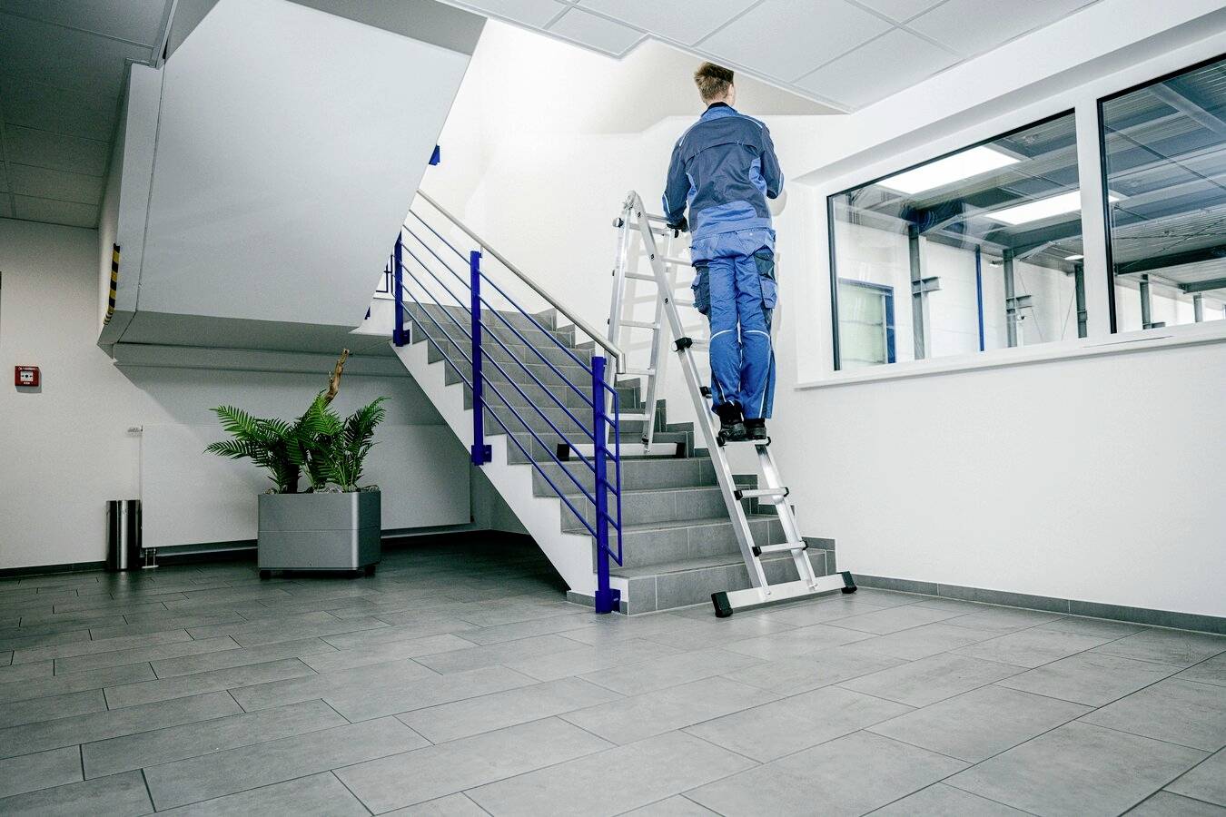 Eine Person steht auf einer Leiter, um eine Wand in einem modernen Bürogebäude zu erreichen. Im Hintergrund sind Treppen und Fenster zu sehen.