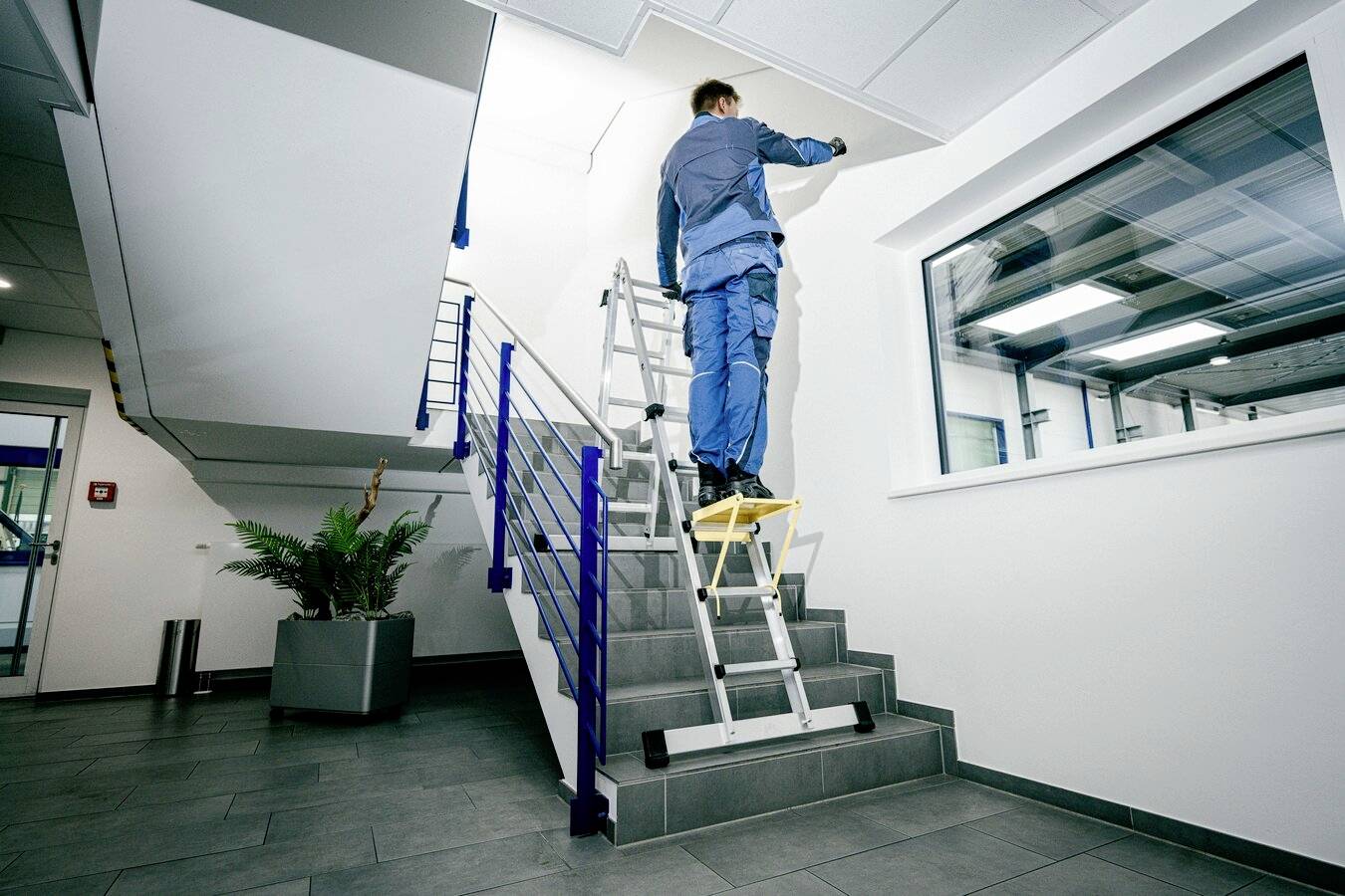 Ein Arbeiter auf einer Leiter streicht die Decke eines Treppenhauses in einem modernen Bürogebäude. Sie sind gut ausgerüstet und tragen Arbeitskleidung.