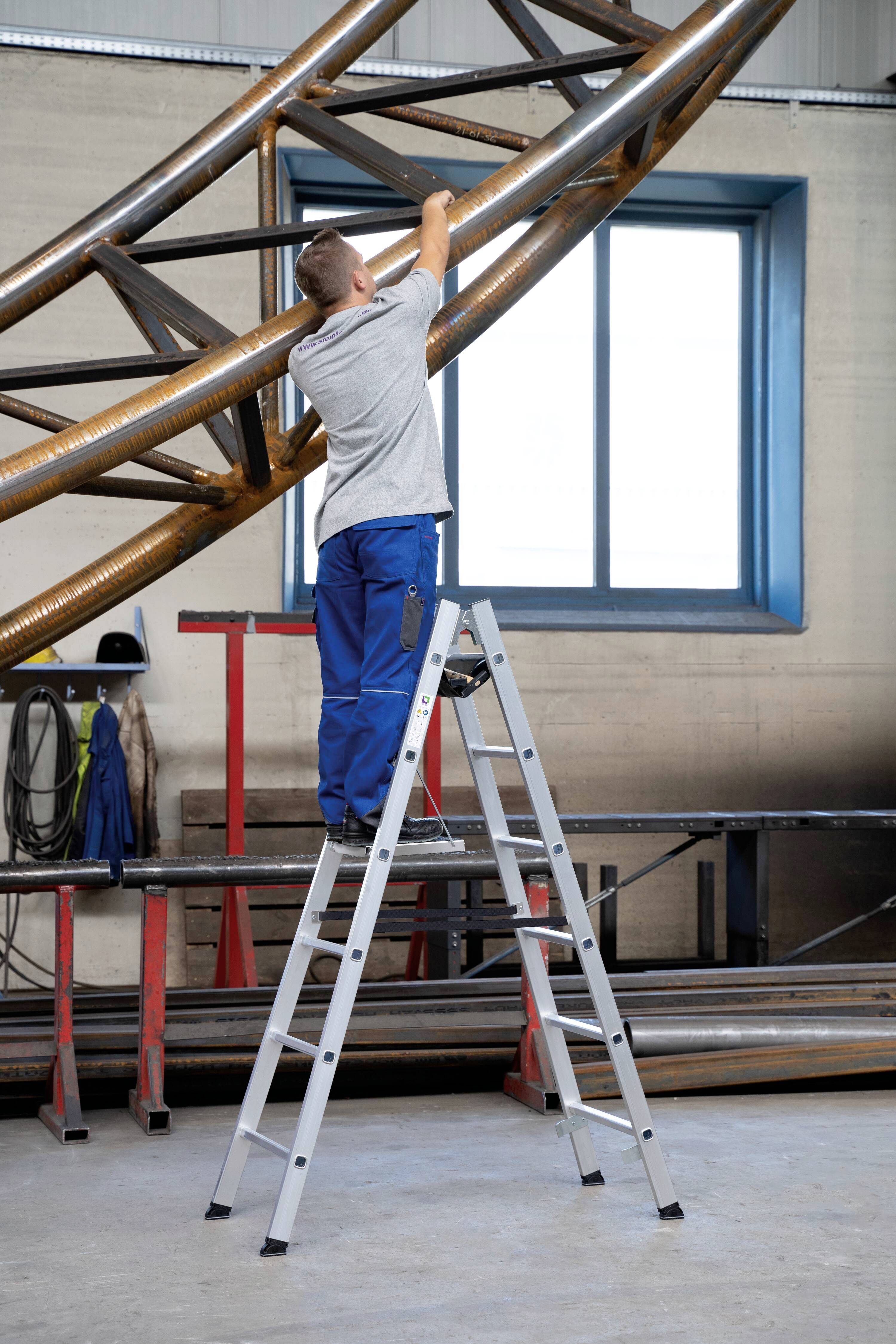 Mann auf Leiter, arbeitet an Metallstruktur in Werkstatt, trägt graues T-Shirt und blaue Hose, Fenster im Hintergrund.