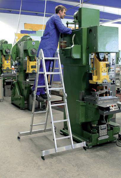 Ein Arbeiter in blauer Arbeitskleidung steht auf einer Leiter und bedient eine große grüne Industriemaschine in einer Fabrikhalle.