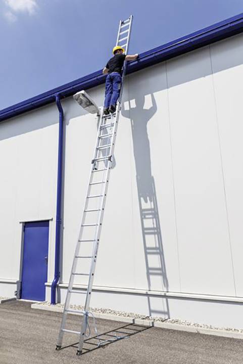 Eine Person klettert mit Helm auf eine lange Leiter, die an einer weißen Wand eines Industriegebäudes lehnt.