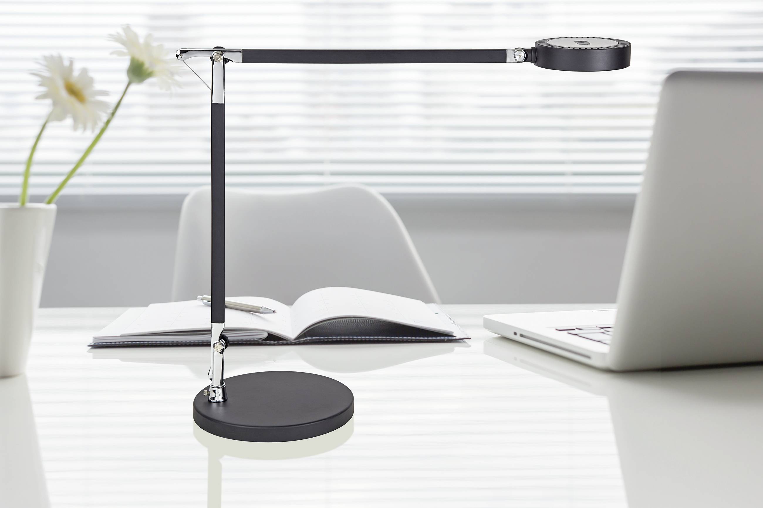 Lichtdurchfluteter Schreibtisch mit einer modernen schwarzen Schreibtischlampe, aufgeklappten Notizbuch und Laptop. Elegante Arbeitsatmosphäre.