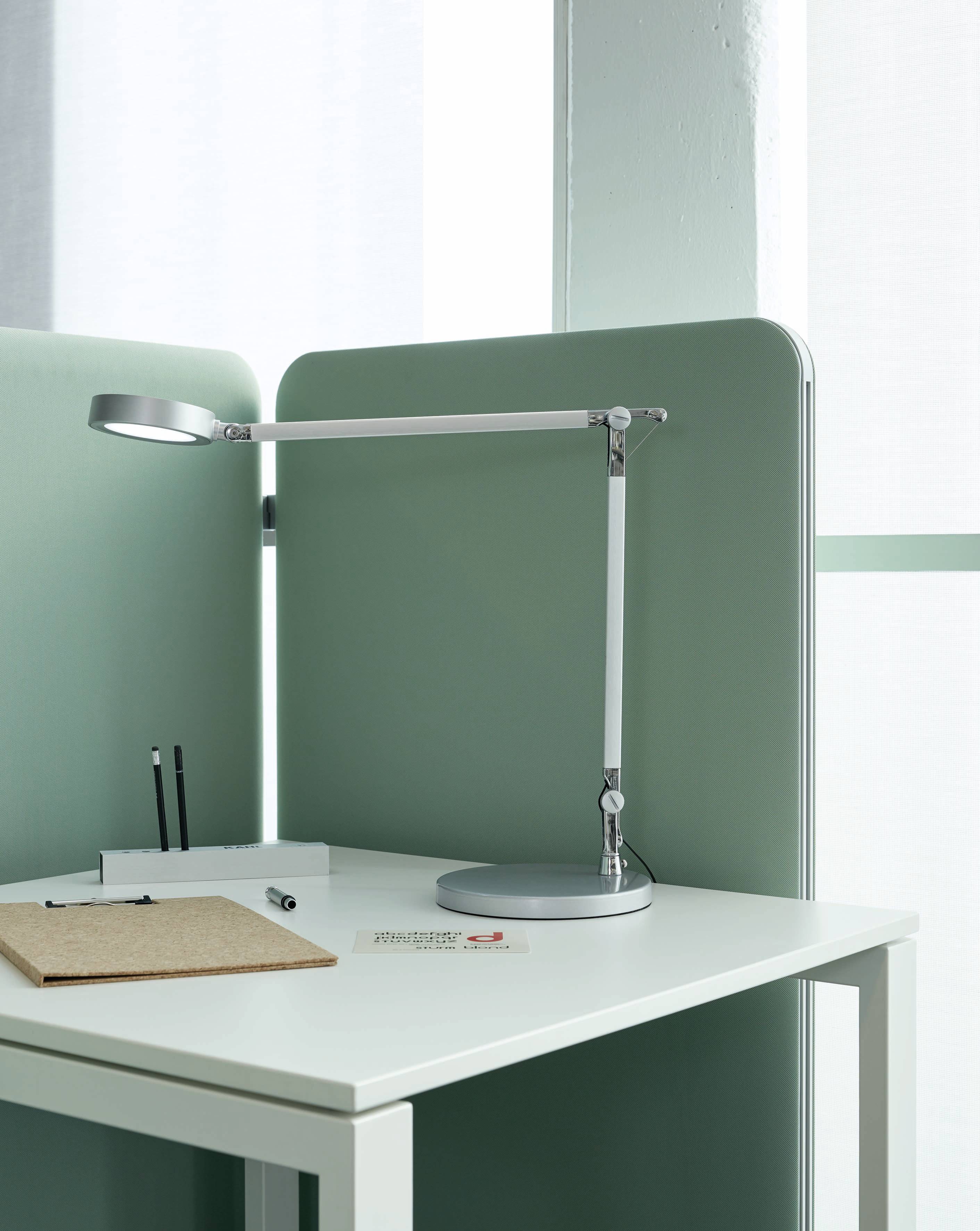 Ein moderner Schreibtisch mit grüner Trennwand, silberner Schreibtischlampe, Stiften, Notizblock und Visitenkarten auf dem weißen Tisch.