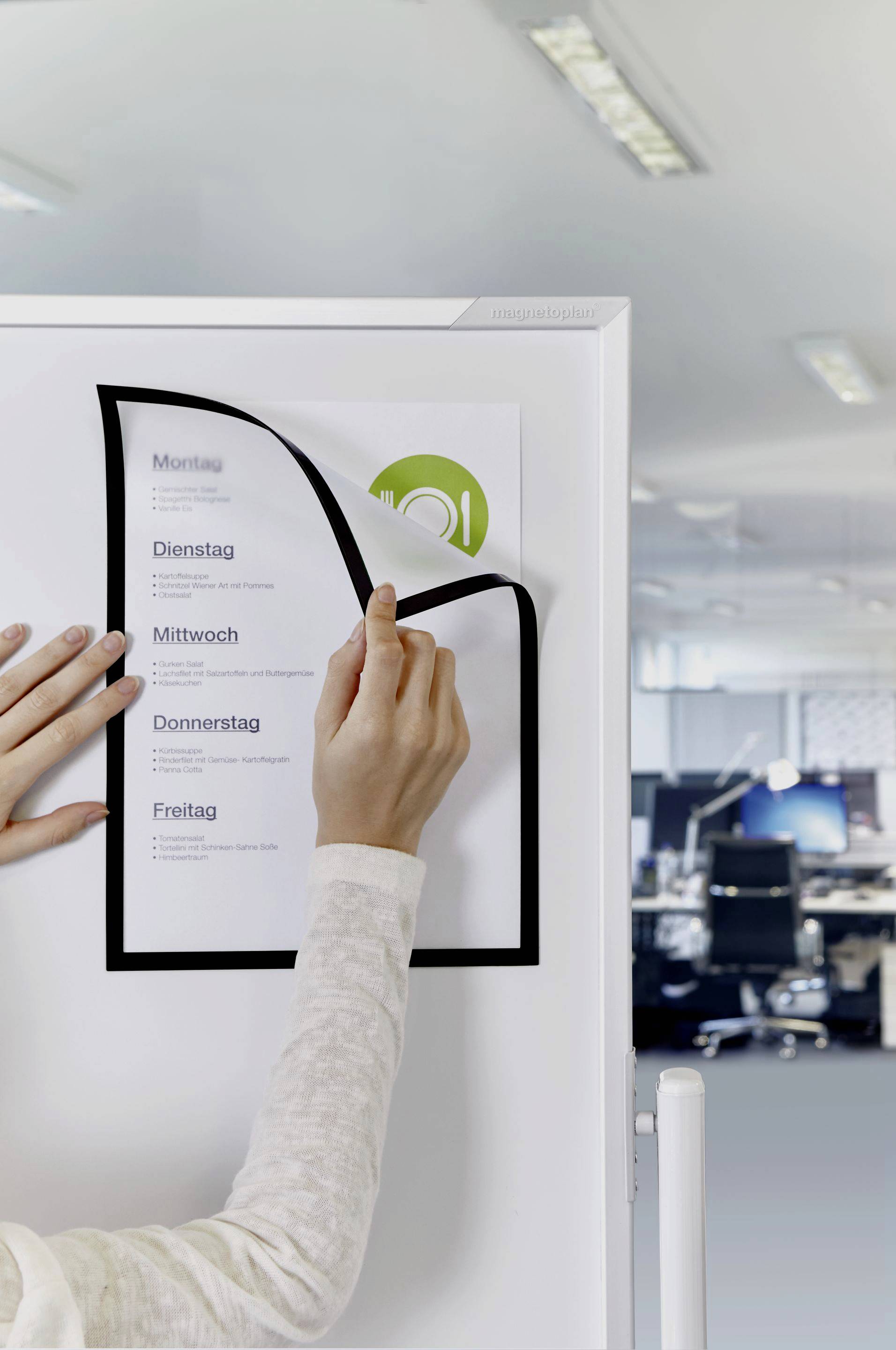 Eine Person klebt ein Blatt mit Wochentagsliste (Montag bis Freitag) an eine Tafel in einem modernen Büro.