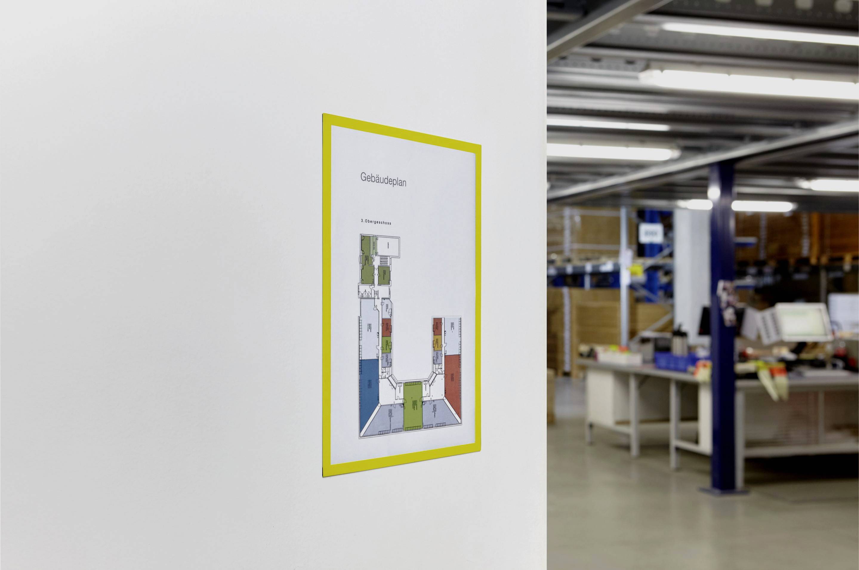 Ein Gebäudeplan hängt an einer weißen Wand in einem Büro. Im Hintergrund sind Regale mit Akten und Schreibtische sichtbar.
