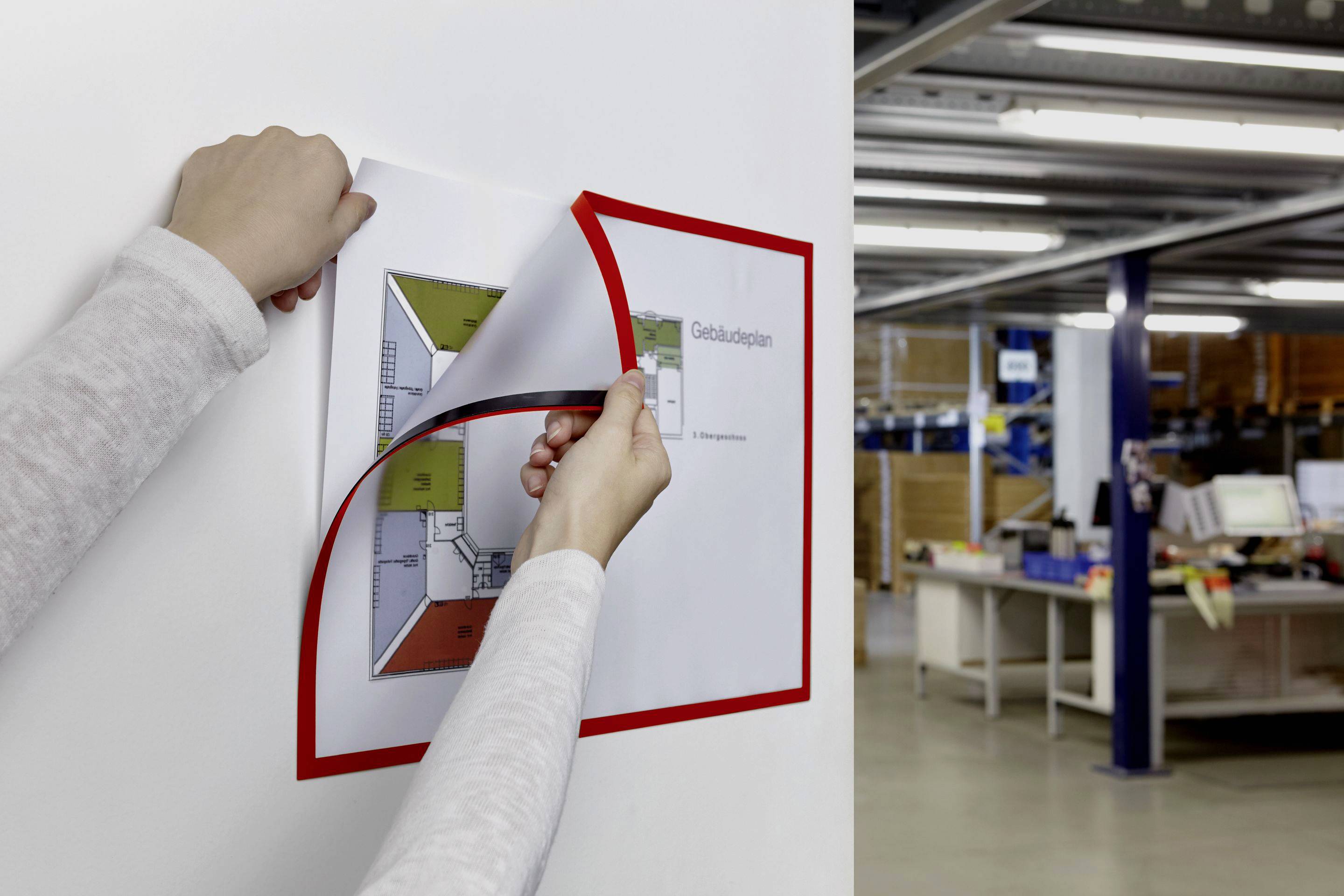 Eine Person hängt einen Gebäudeplan an die Wand in einem Büro auf. Im Hintergrund sind Regale und Schreibtische zu sehen.