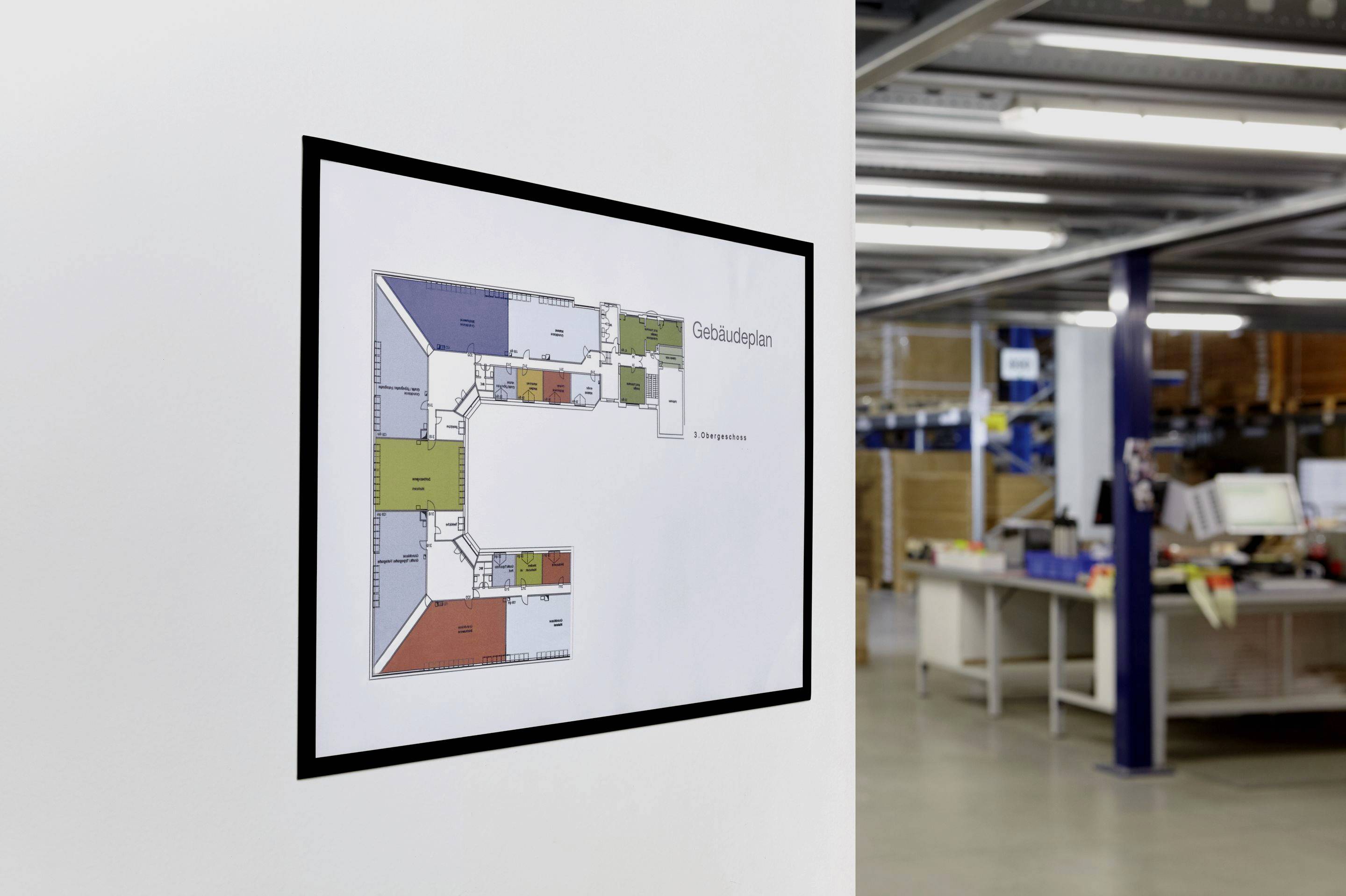 Gebäudeplan an der Wand eines Büros. Zeigt verschiedene farbig kodierte Räume und Flure. Büroumgebung im Hintergrund.