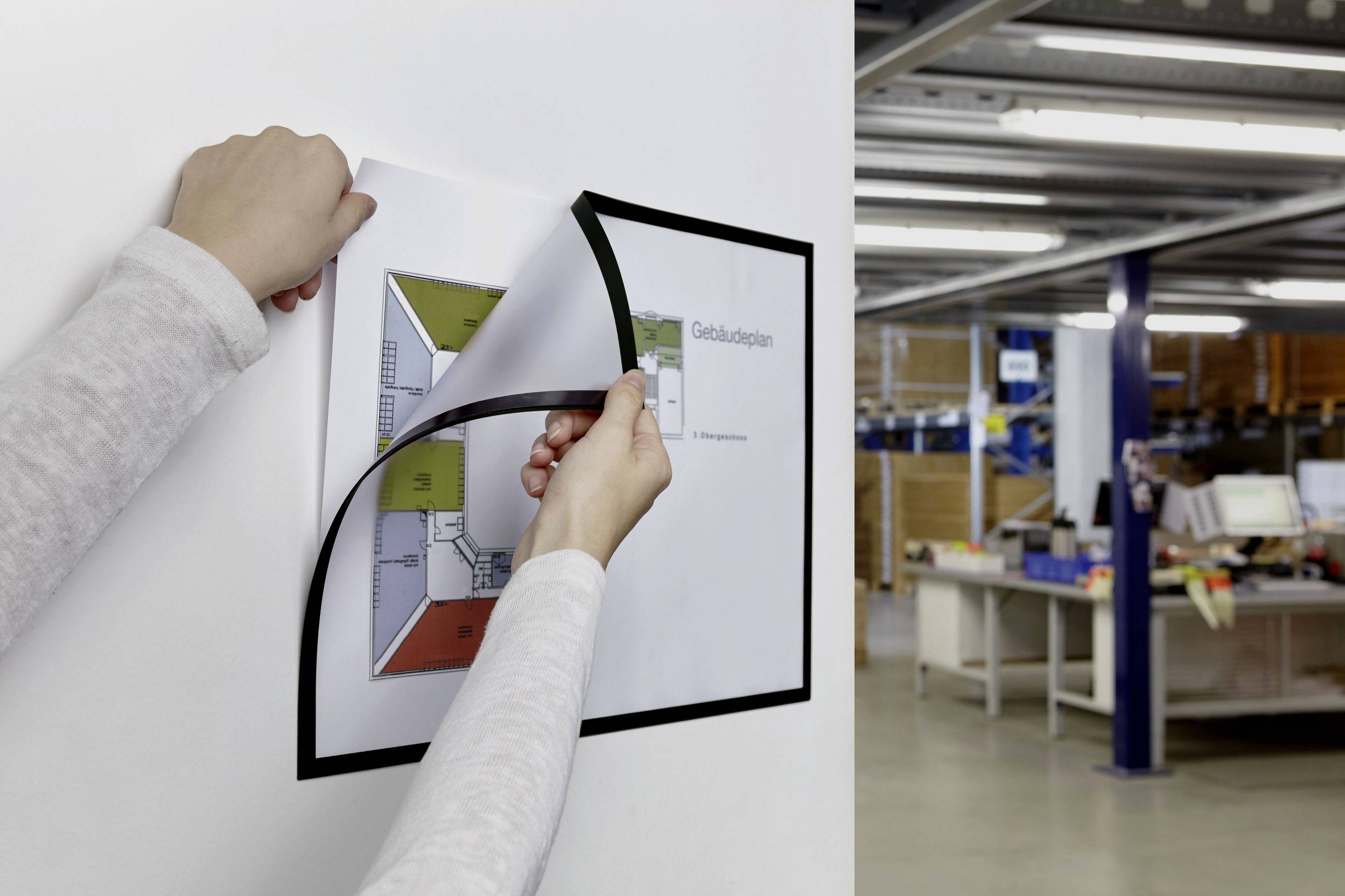 Eine Person hängt einen Gebäudeplan an einer Wand auf. Im Hintergrund sind Regale und ein Schreibtisch in einem Büro zu sehen.