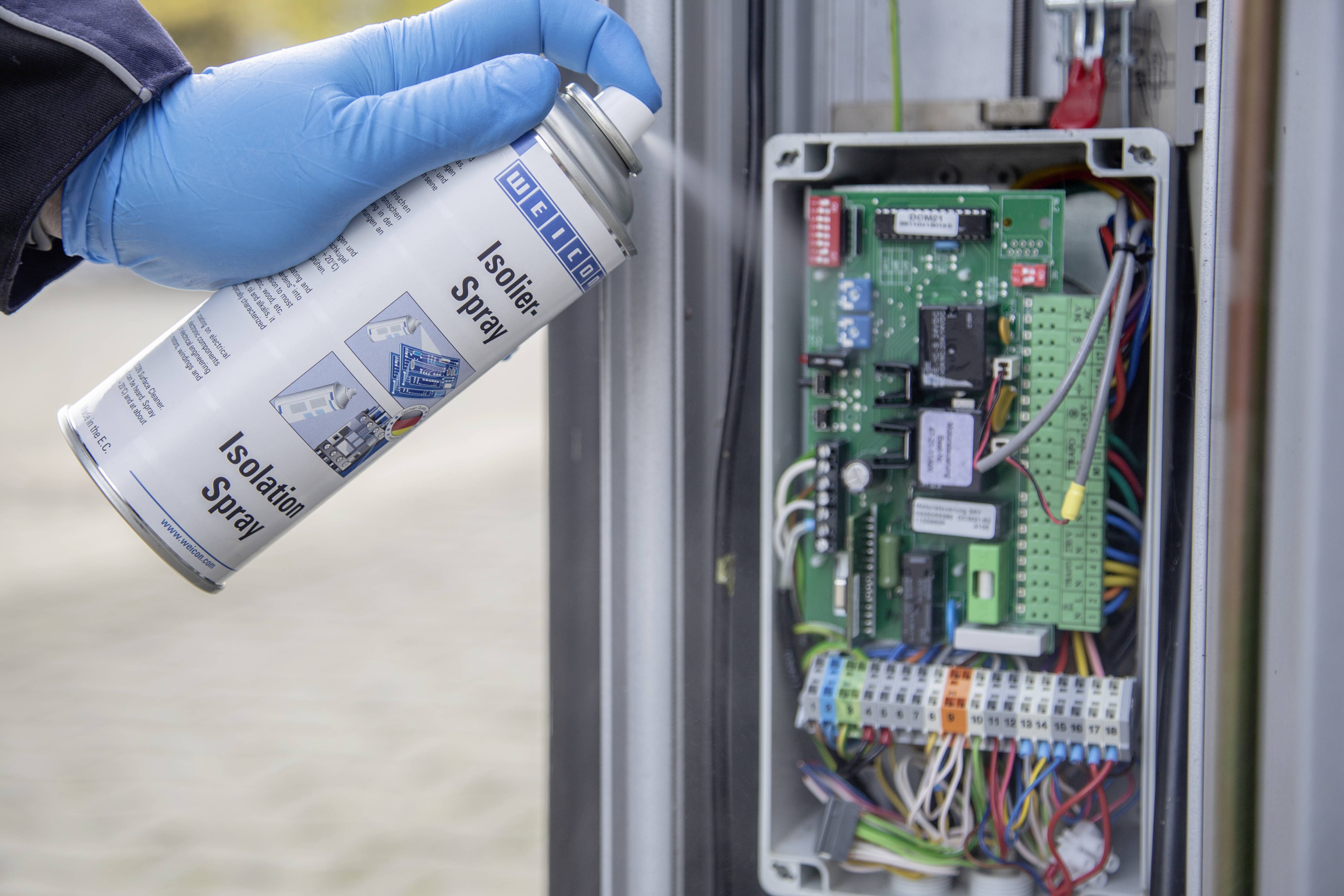 Eine Person sprüht Isolierspray auf eine geöffnete elektronische Schaltbox mit vielen Kabeln und Komponenten.