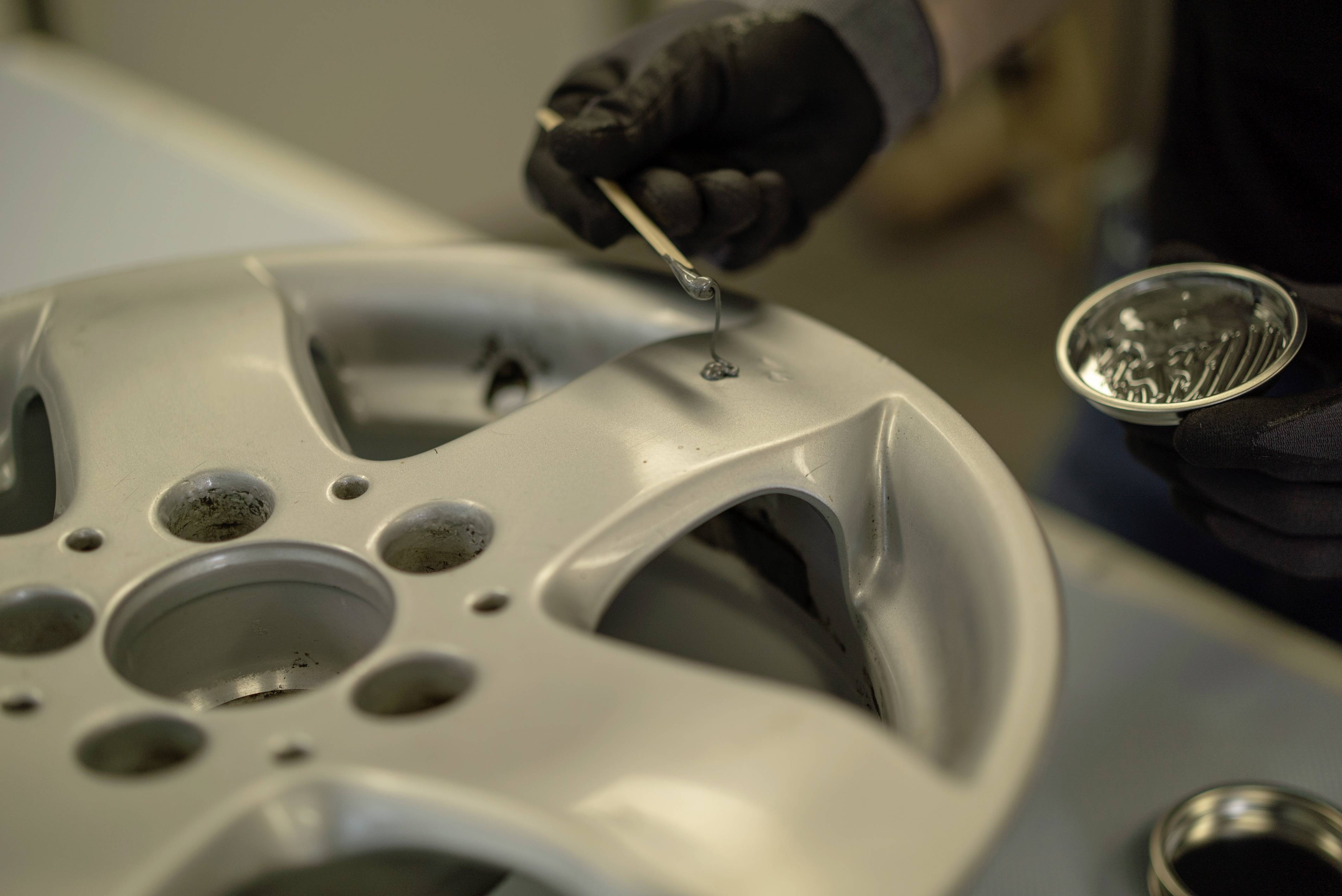 Eine Person repariert eine silberne Autoradfelge, trägt mit einem kleinen Werkzeug eine Substanz auf, um Kratzer auszubessern.