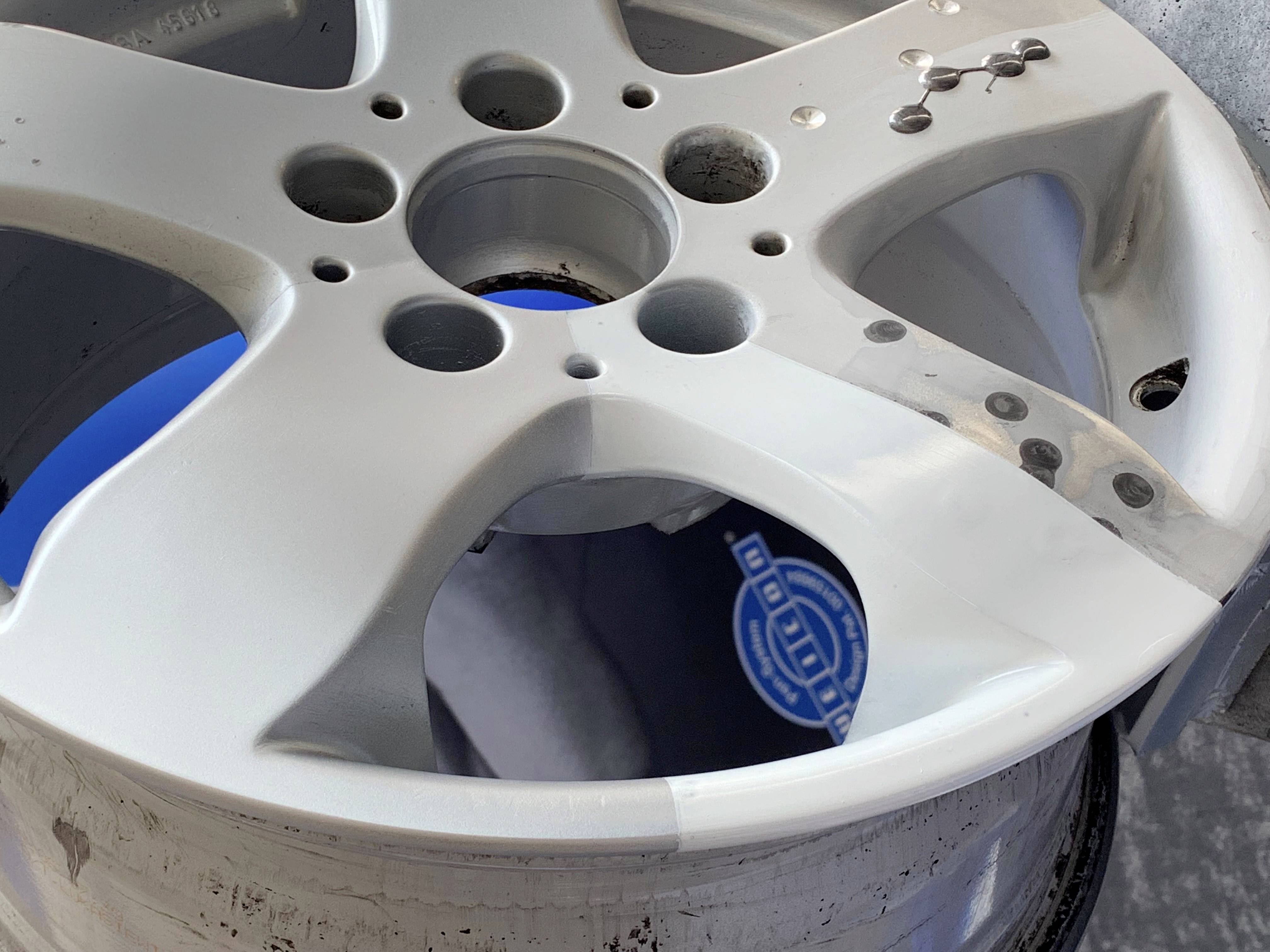 Abgenutzte silberne Autoreifenfelge mit mehreren Löchern und Kratzspuren. Im Hintergrund ein unscharfes blaues Element sichtbar.