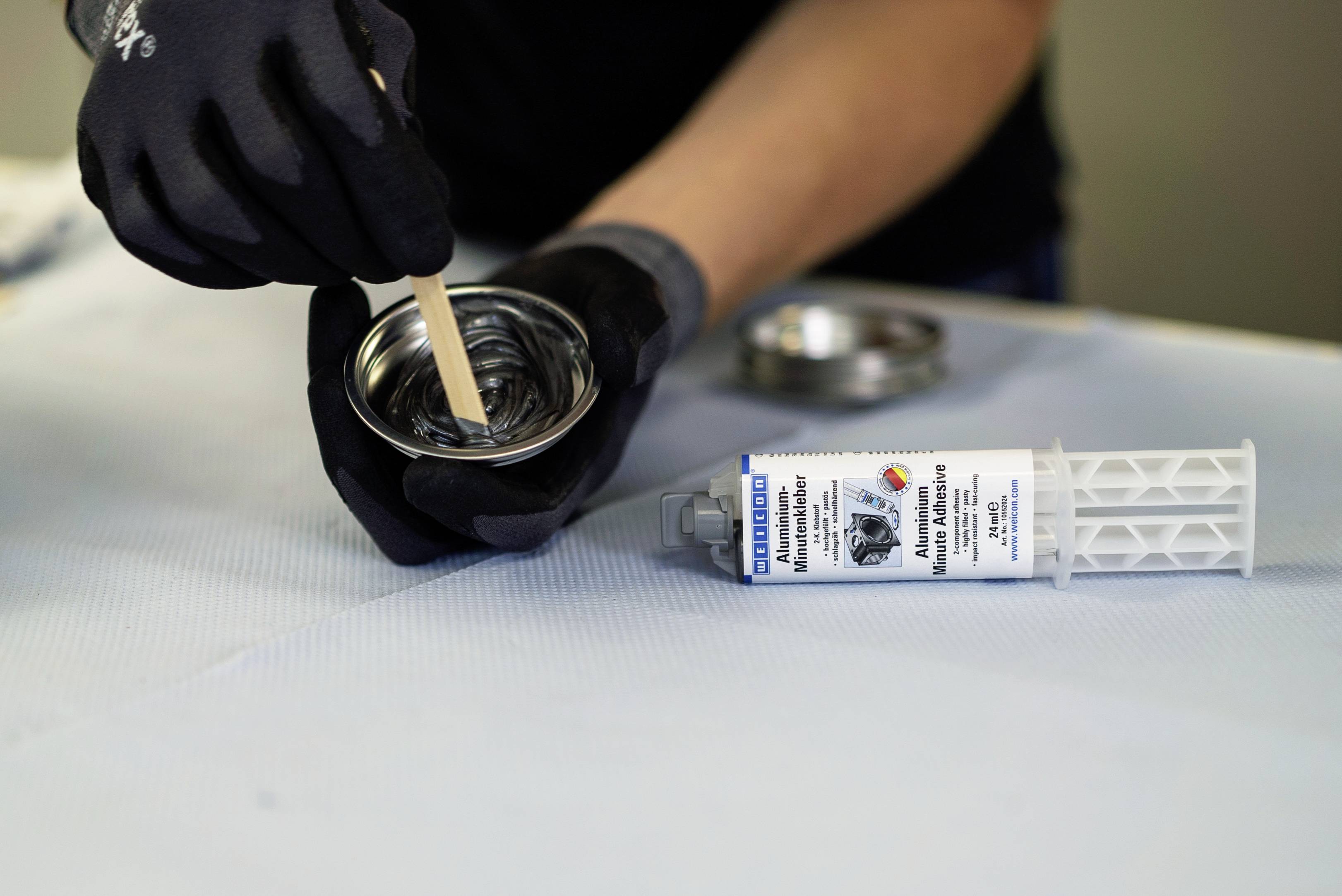Person mit Handschuhen mischt Inhalt in einer Metallschale. Eine Tube Kleber liegt daneben auf dem Tisch.