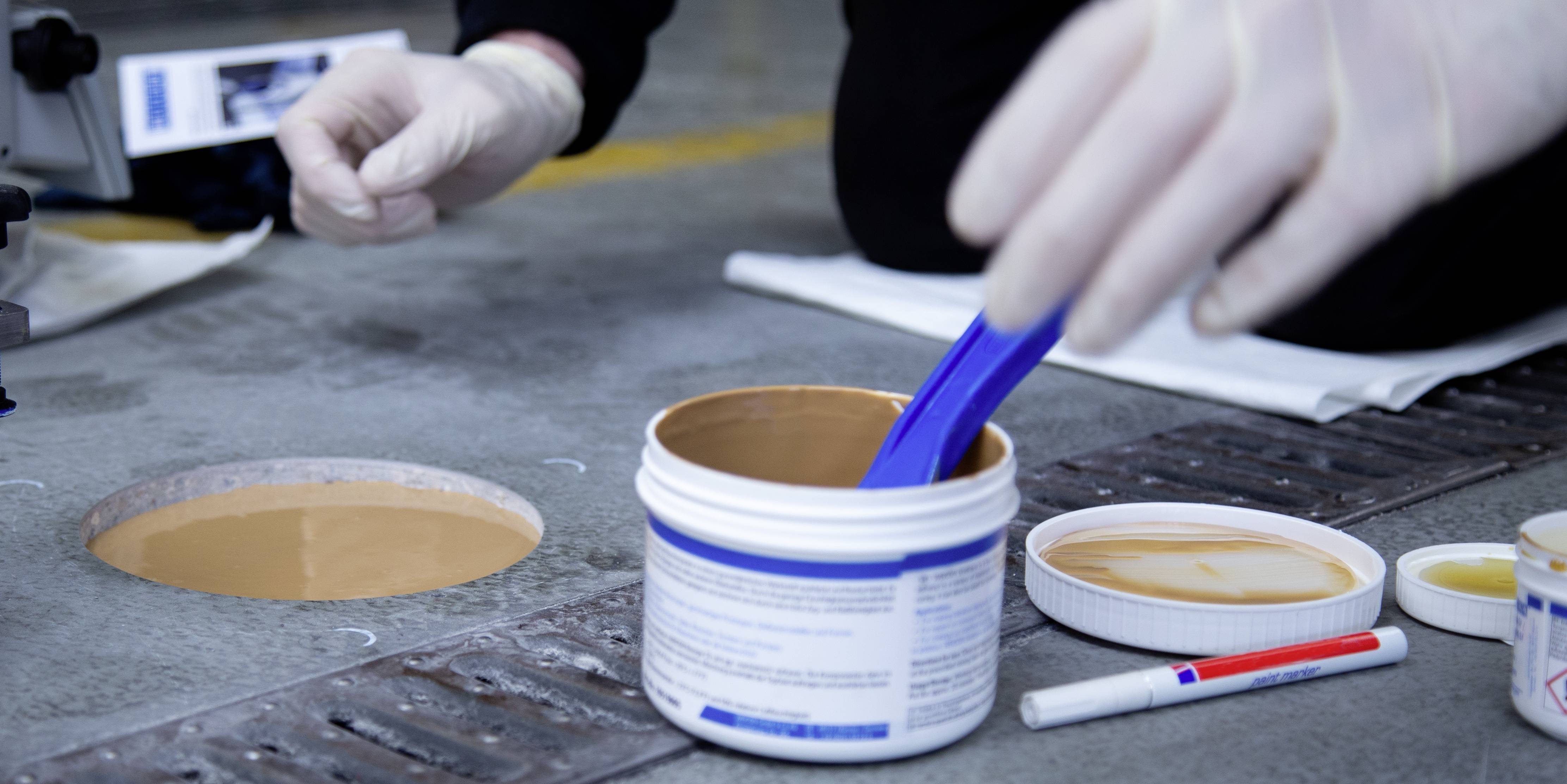 Person trägt mit einem blauen Spatel eine Substanz aus einer Dose auf eine Betonoberfläche auf. Person trägt weiße Handschuhe.