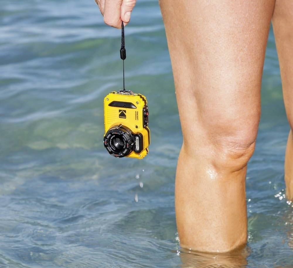 Eine gelbe wasserdichte Kamera wird in der Hand einer Person gehalten, die bis zu den Knien im klaren Wasser steht.