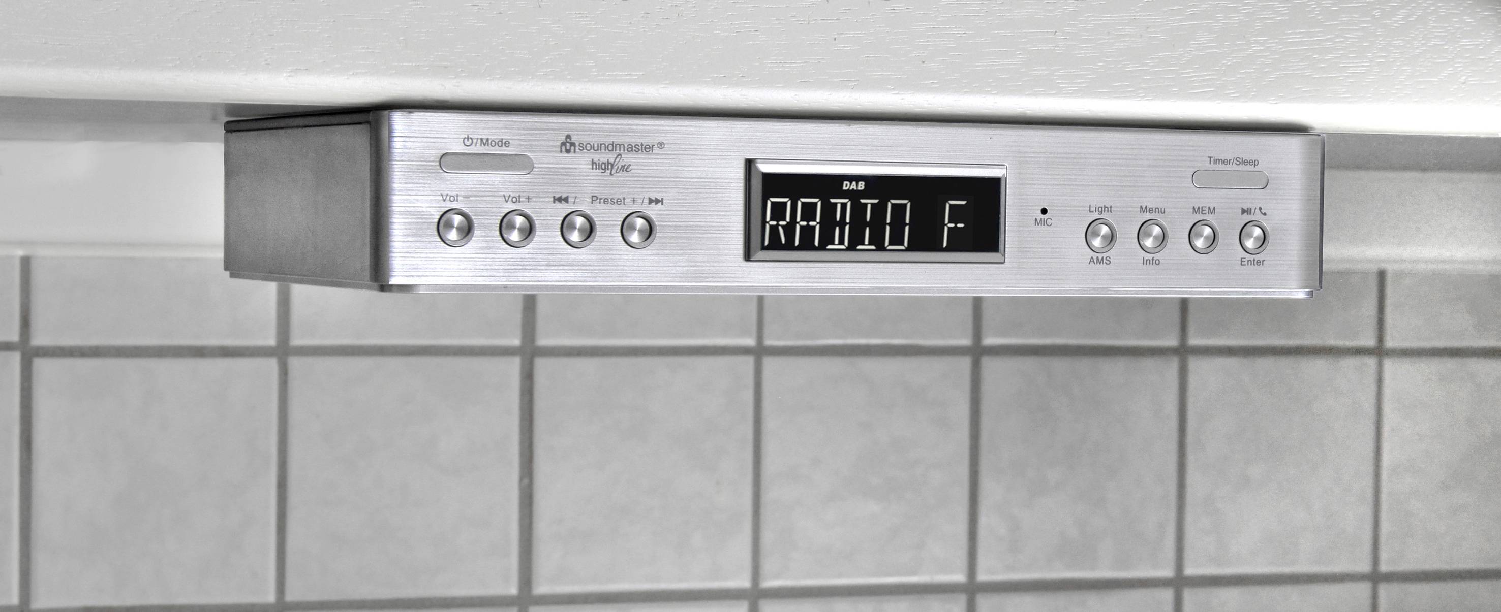 Radio unter Küchenregal montiert, zeigt 'RADIO F' auf Display, mit Tasten für Sender, Lautstärke und Funktionen. Hintergrund: graue Fliesen.
